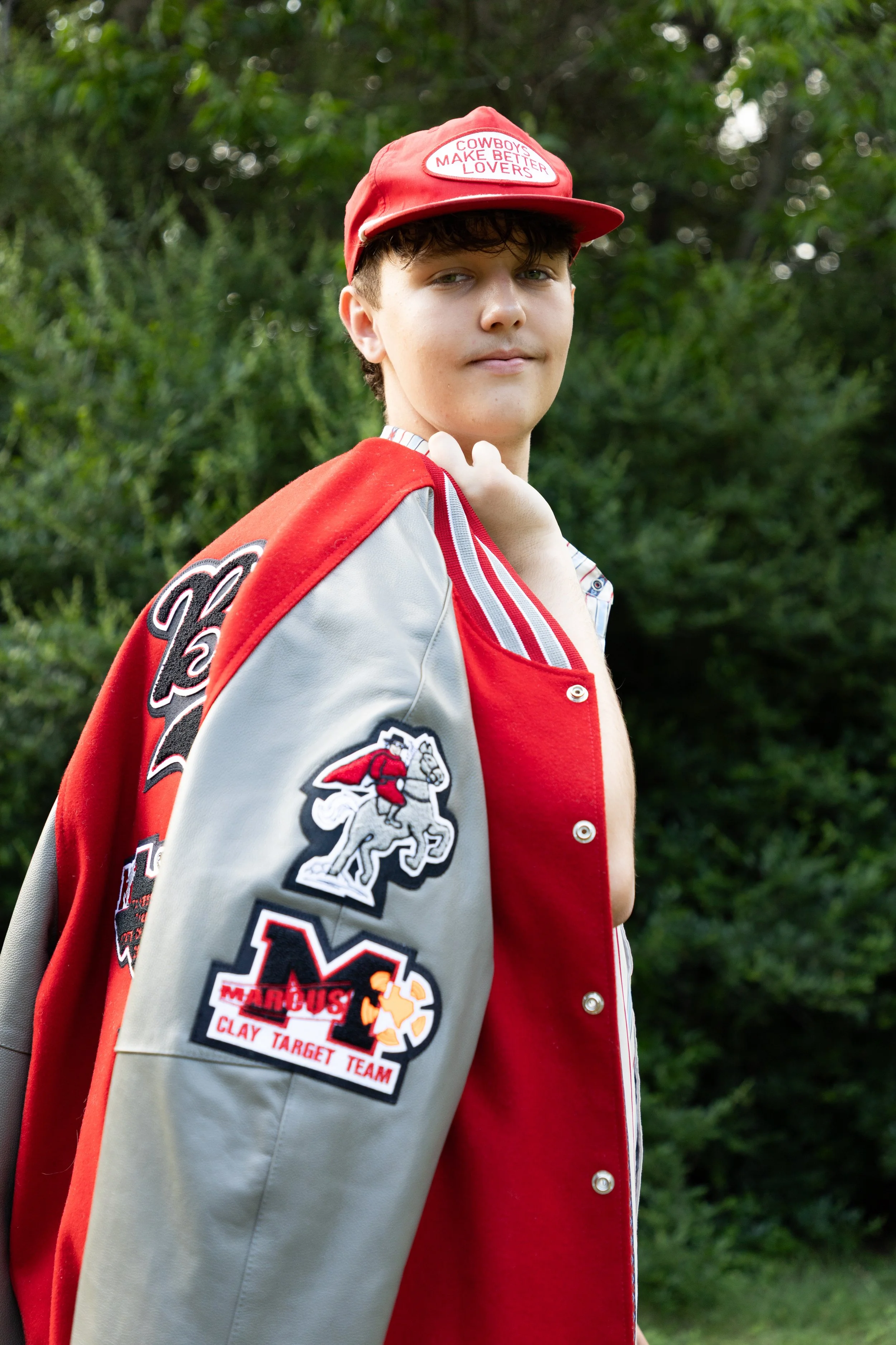 A young man outdoors wearing a red and gray letterman jacket with patches, a red cap with a patch that reads 'Cowboys Make Better Lovers,' and a striped shirt underneath, standing in front of green foliage.