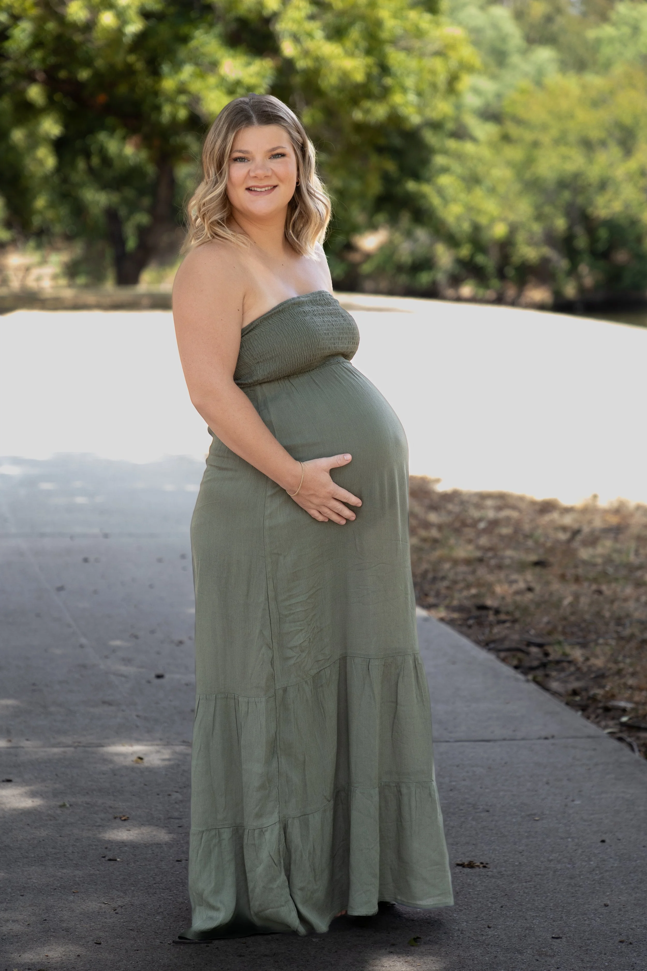 A pregnant woman in a strapless green maxi dress standing outdoors on a paved path, holding her belly with one hand, smiling, with trees and sunlight in the background.
