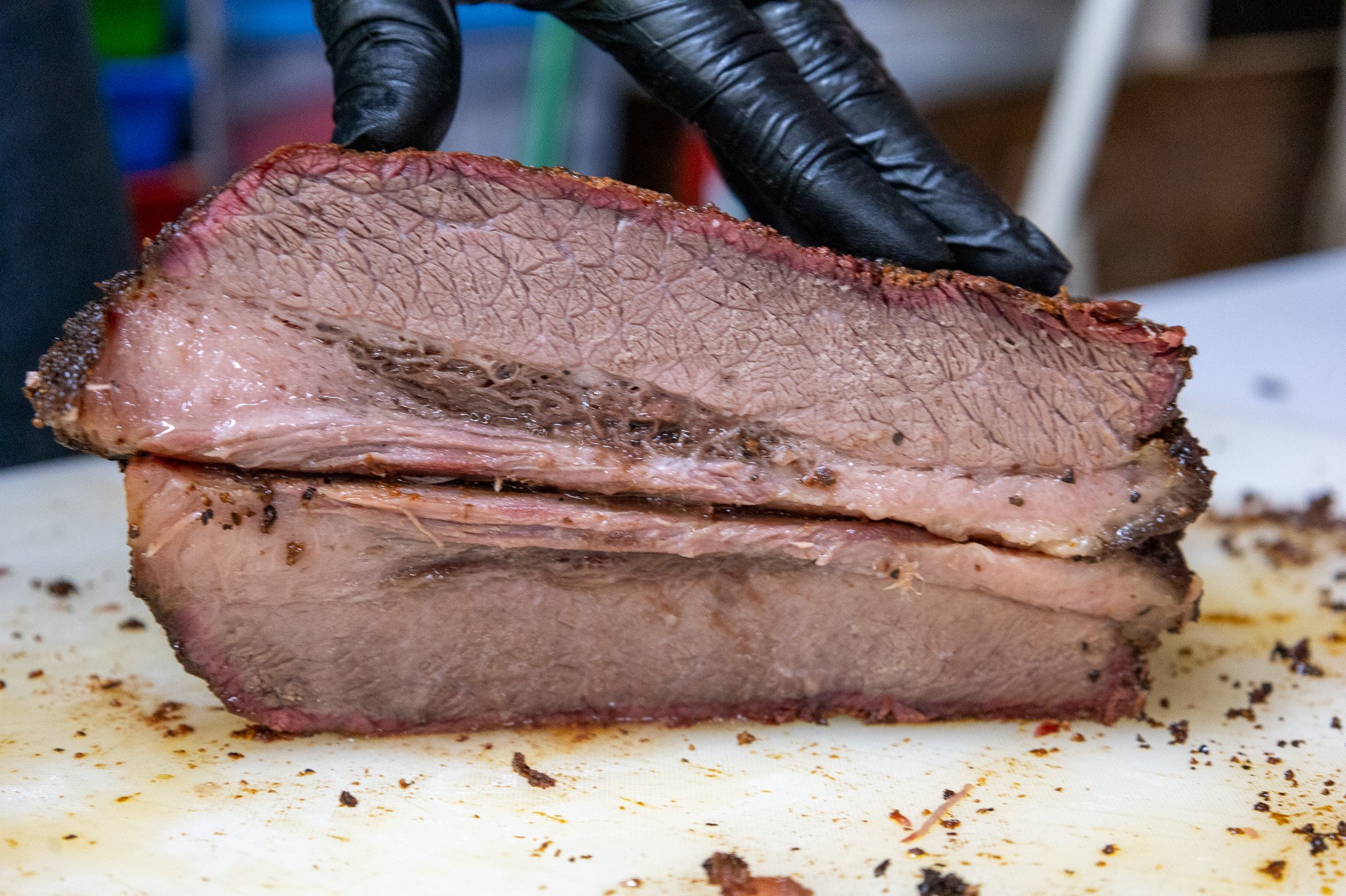 A person wearing black gloves slicing a cooked brisket on a white cutting board with seasoning crumbs