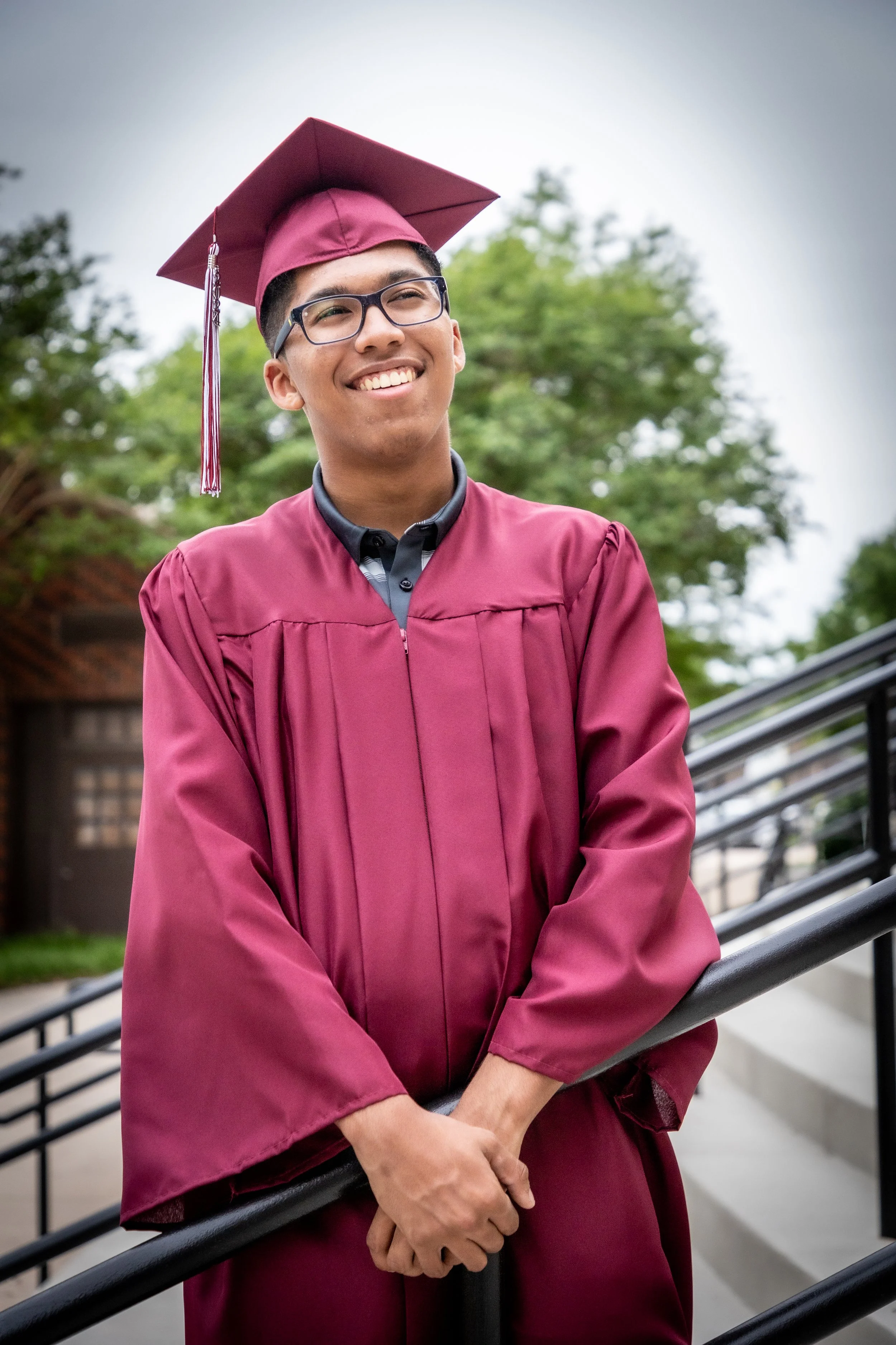 Young man in maroon graduation cap and gown smiling outdoors on steps.