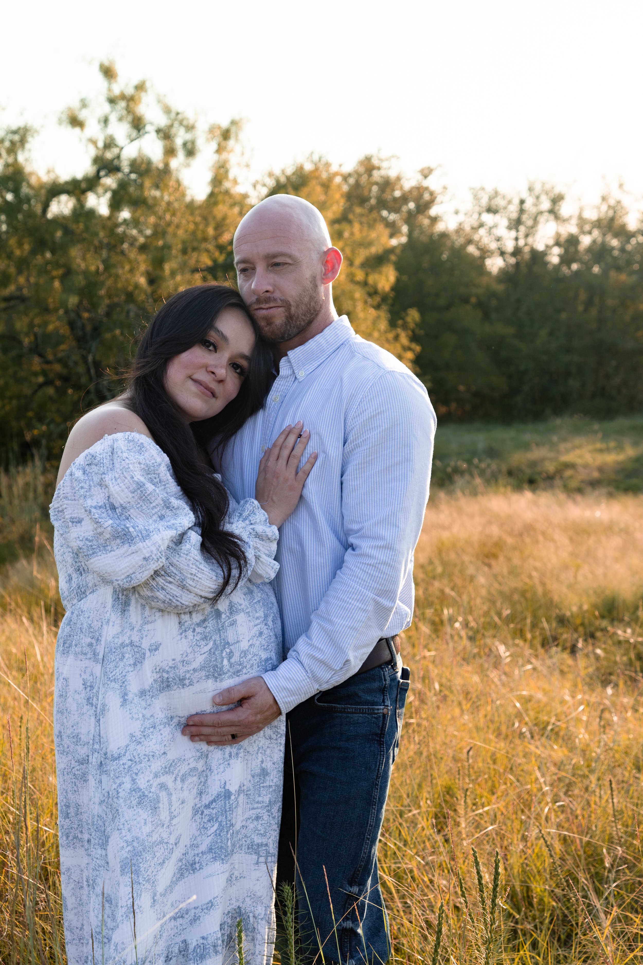 A couple standing in a grassy field during sunset, with the woman resting her head on the man's shoulder and the man gently holding her belly, both looking peaceful.