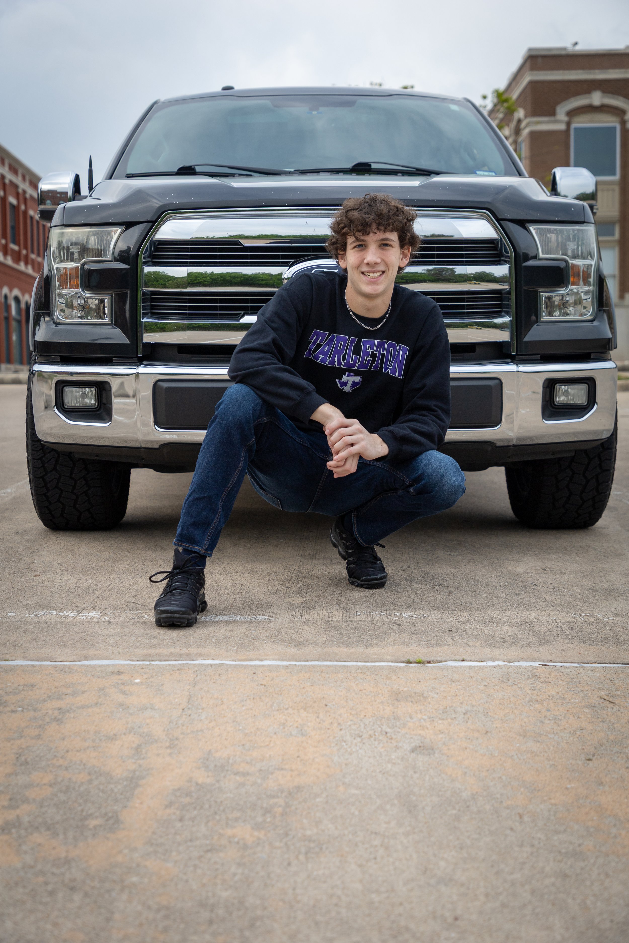 A young man with curly hair squatting in front of a large black and chrome pickup truck, smiling at the camera. He is wearing a black sweatshirt with purple and white text and dark jeans, and is outdoors in front of a brick building.