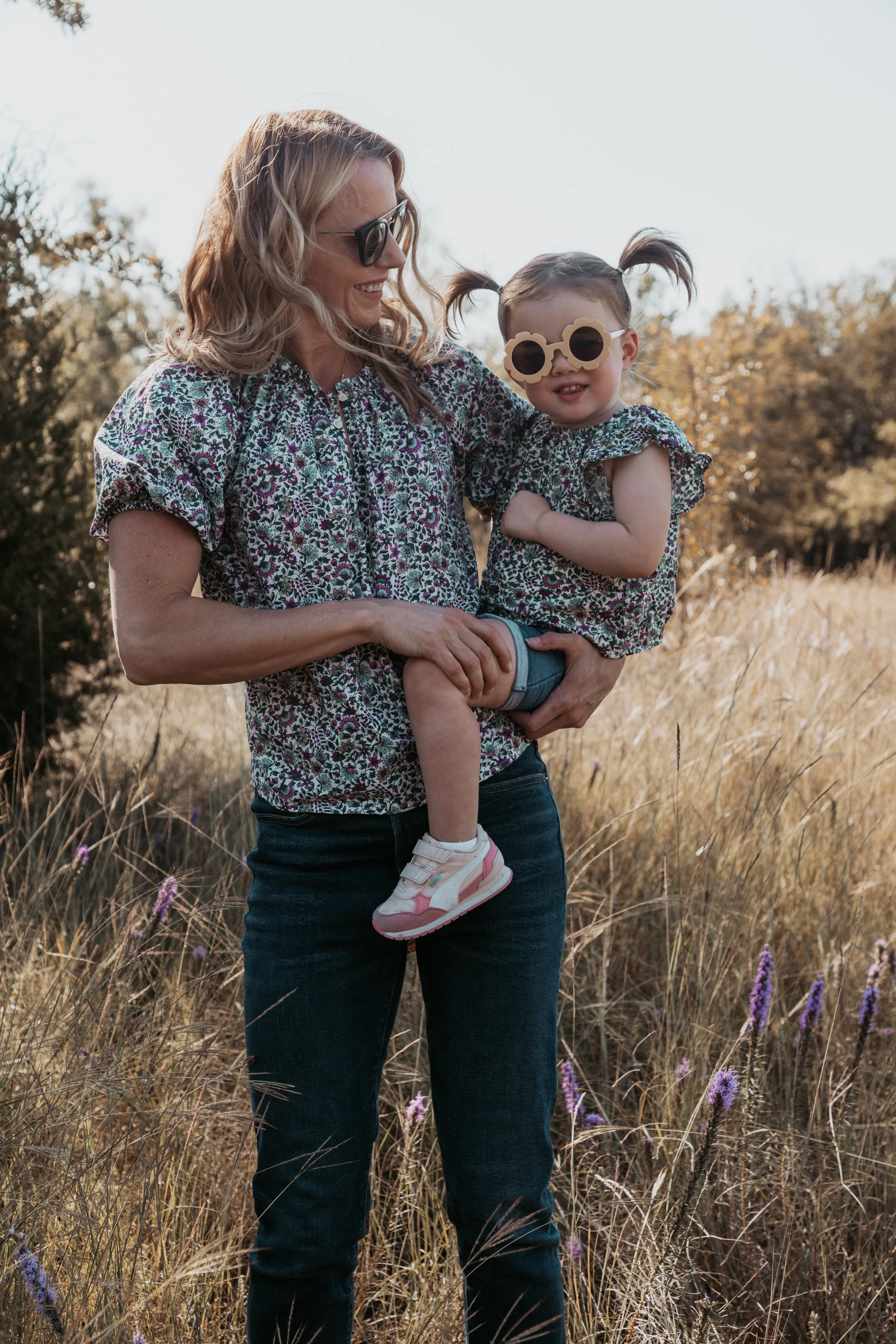 A woman carrying a young girl in a field of tall grass and wildflowers, both wearing matching floral tops, with the girl wearing sunglasses and playful shoes.