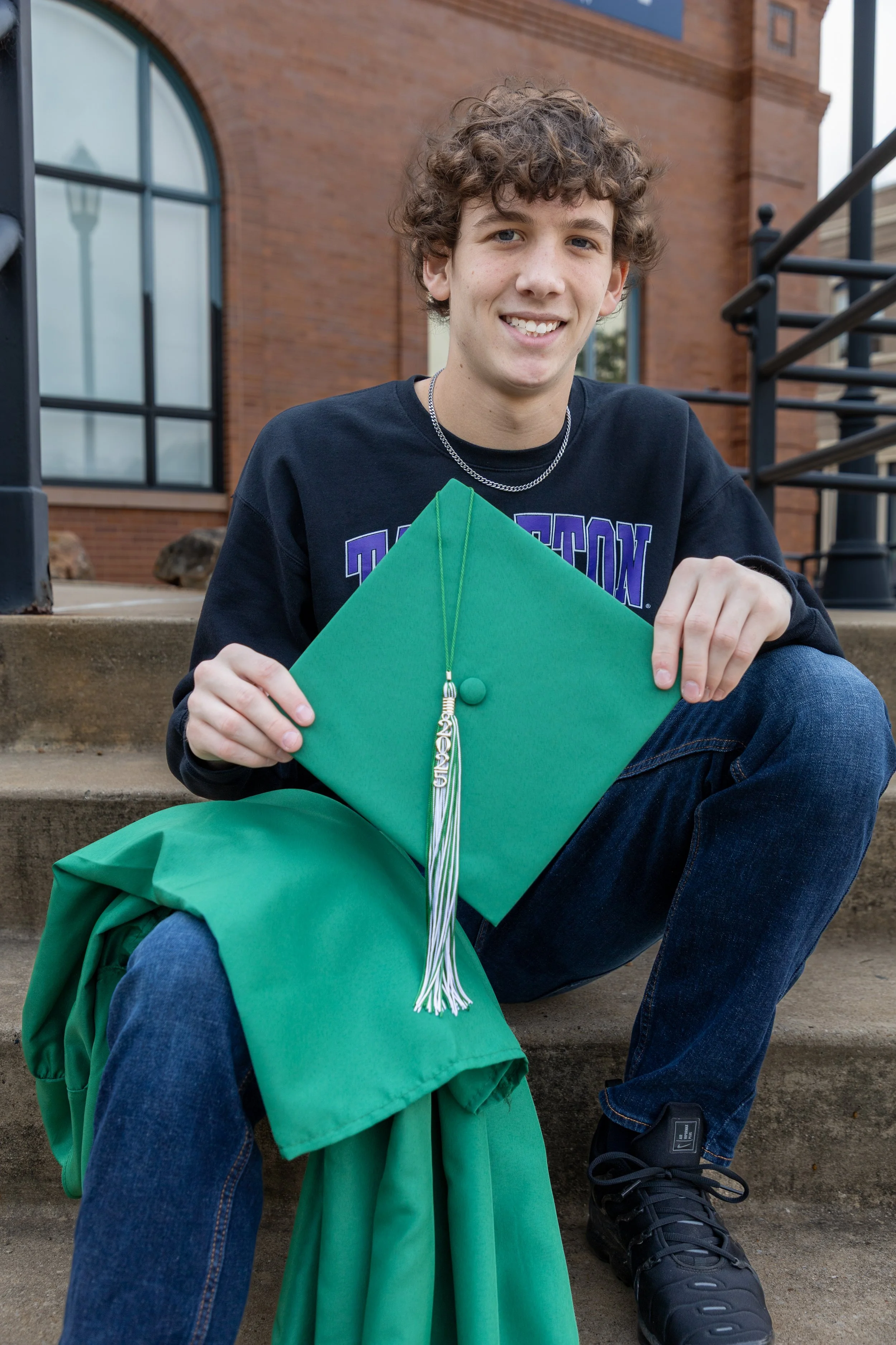 A young man with curly hair sitting on steps outside a brick building, holding a green graduation cap decorated with a silver tassel.