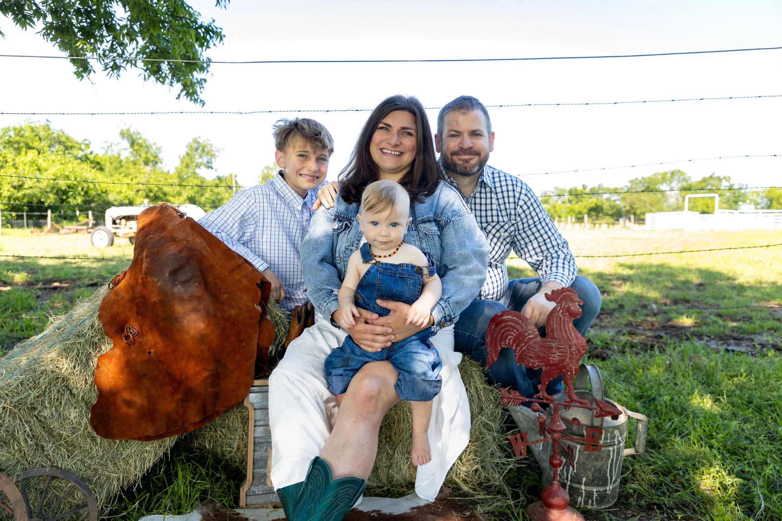 A family of four sitting outdoors on a farm, smiling at the camera, with hay bales, farm tools, and greenery in the background.