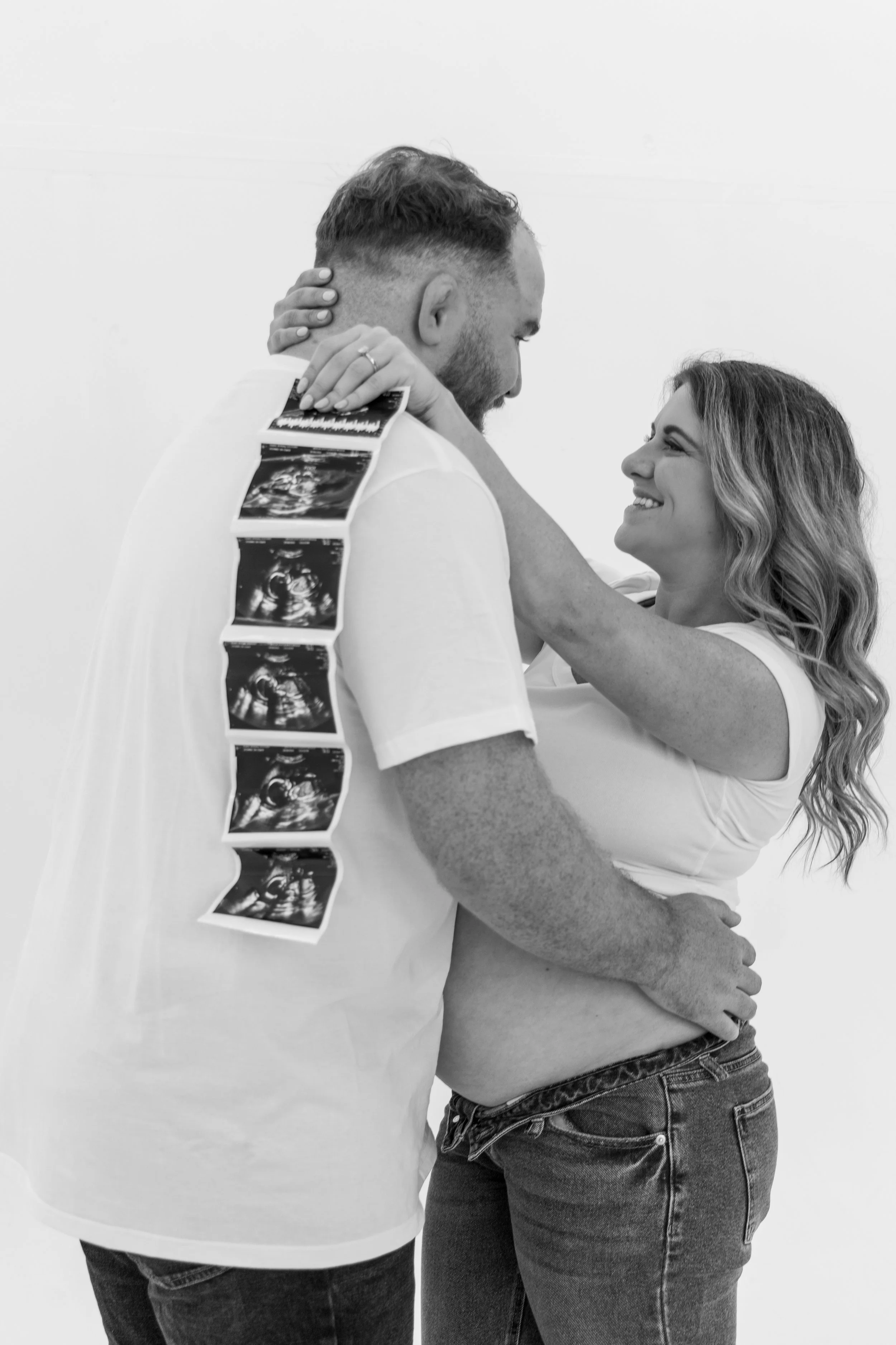 A couple holding each other closely, smiling and looking into each other's eyes in a black and white photo. The woman has wavy hair and is wearing jeans, and the man has a short beard and is holding ultrasound images.