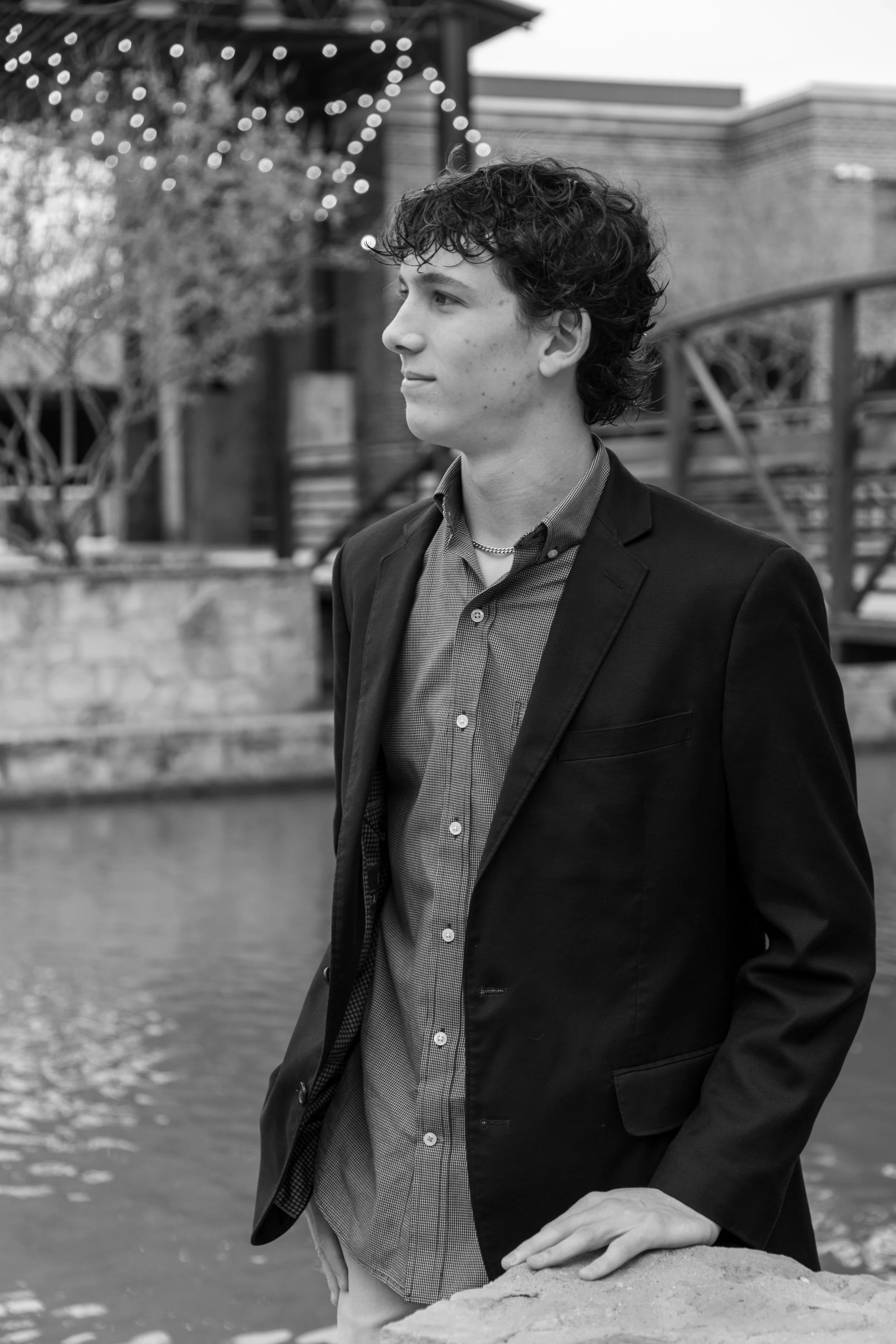A young man with curly hair dressed in a dark blazer and a button-up shirt, standing outdoors by a body of water, with a background of buildings, trees, and string lights.