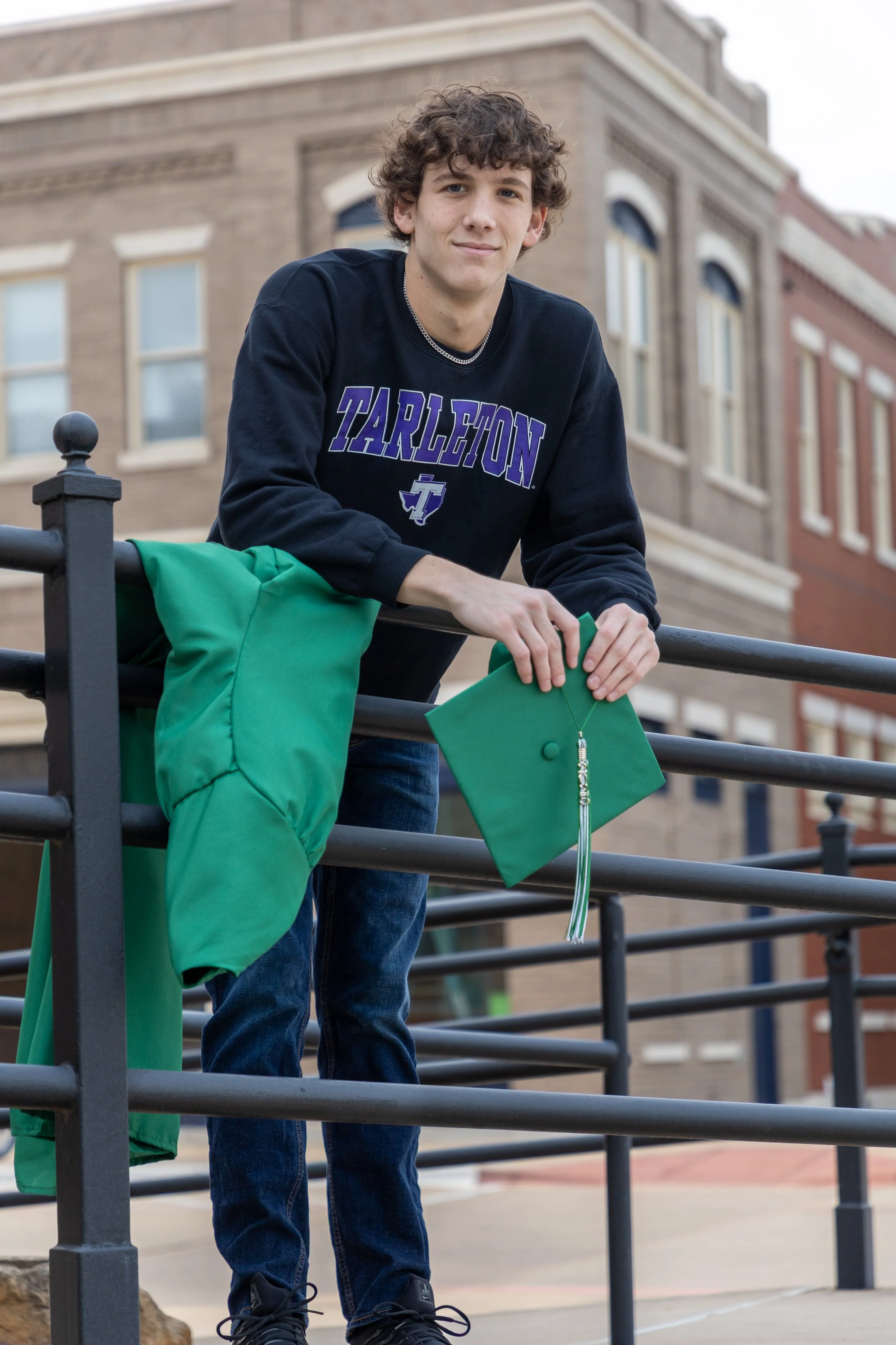 A young man standing outdoors, holding a green graduation cap, with a green graduation gown hanging on a black railing, in front of a brick building.