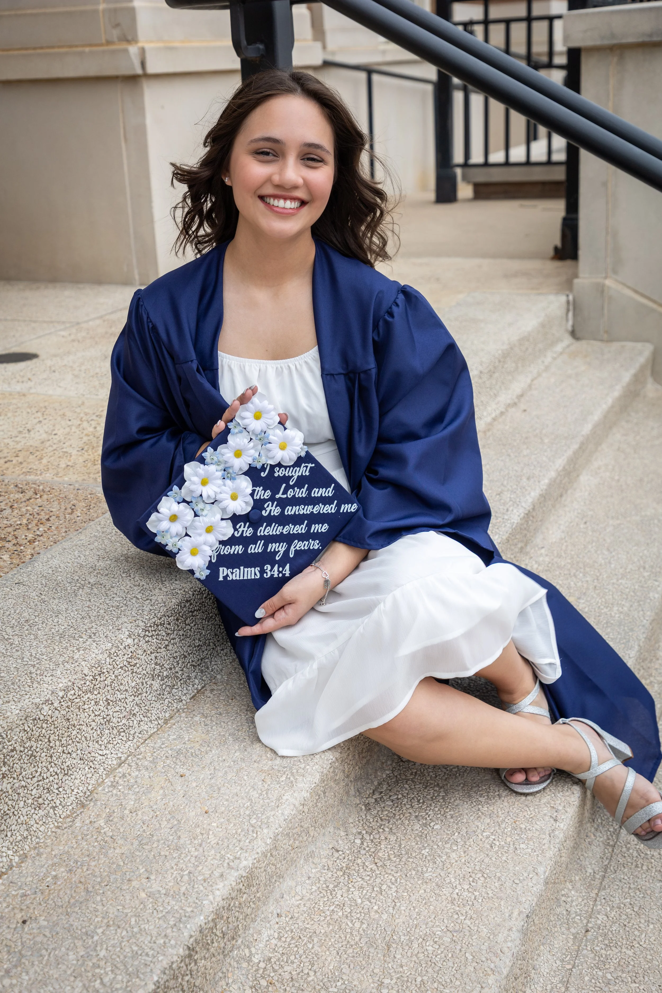 A young woman sitting on outdoor stairs wearing a blue graduation gown and holding a floral diploma cover with a Psalms 34:4 quote and daisies attached.