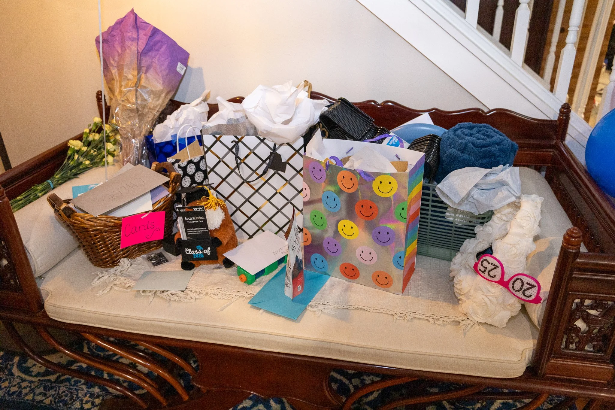 A wooden bench filled with various gifts, including flowers, gift bags, cards, and tissue paper, with a staircase in the background.