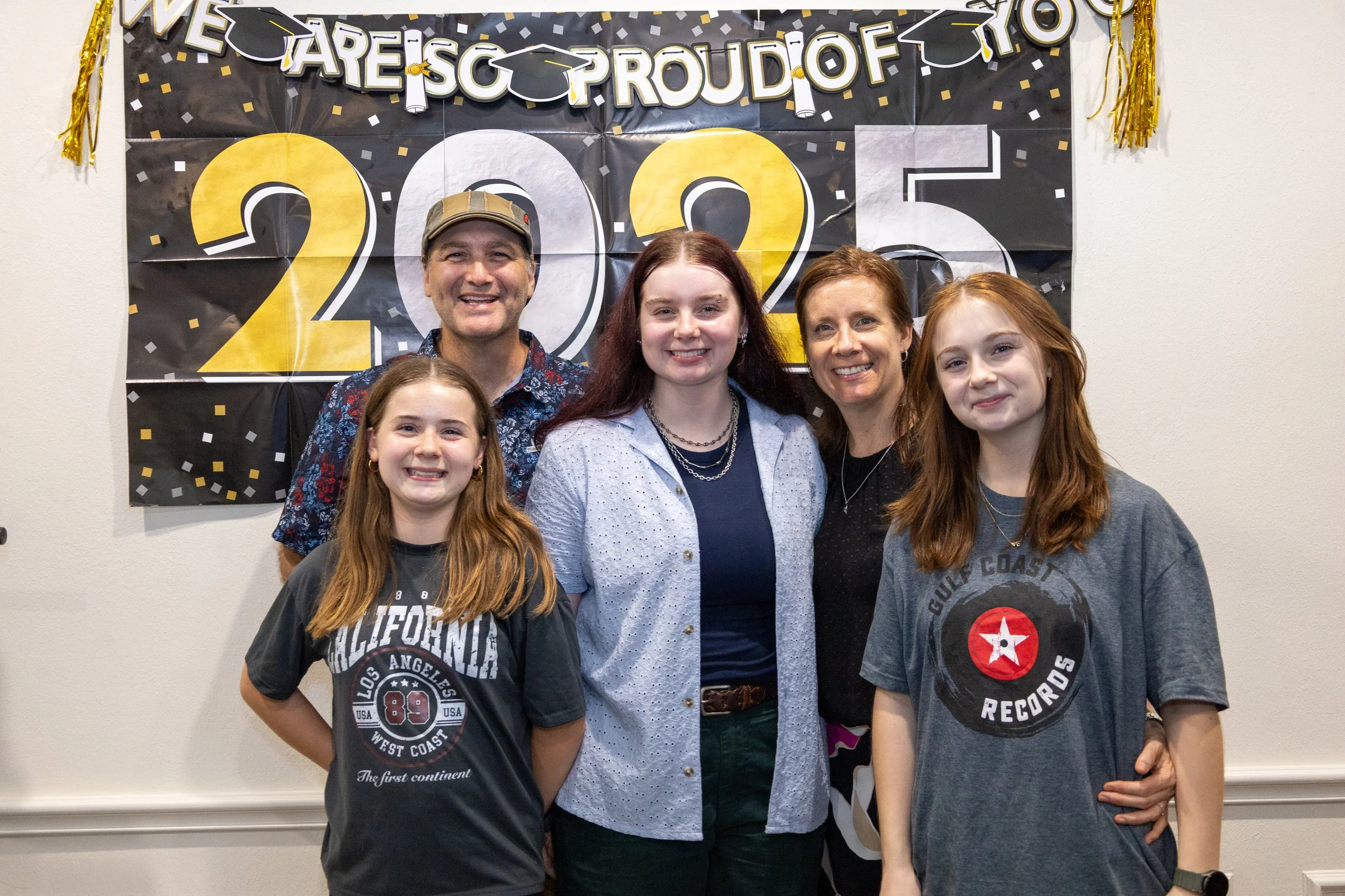 A group of five people, two men and three women, standing in front of a black and gold graduation celebration banner. They are smiling for the photo.