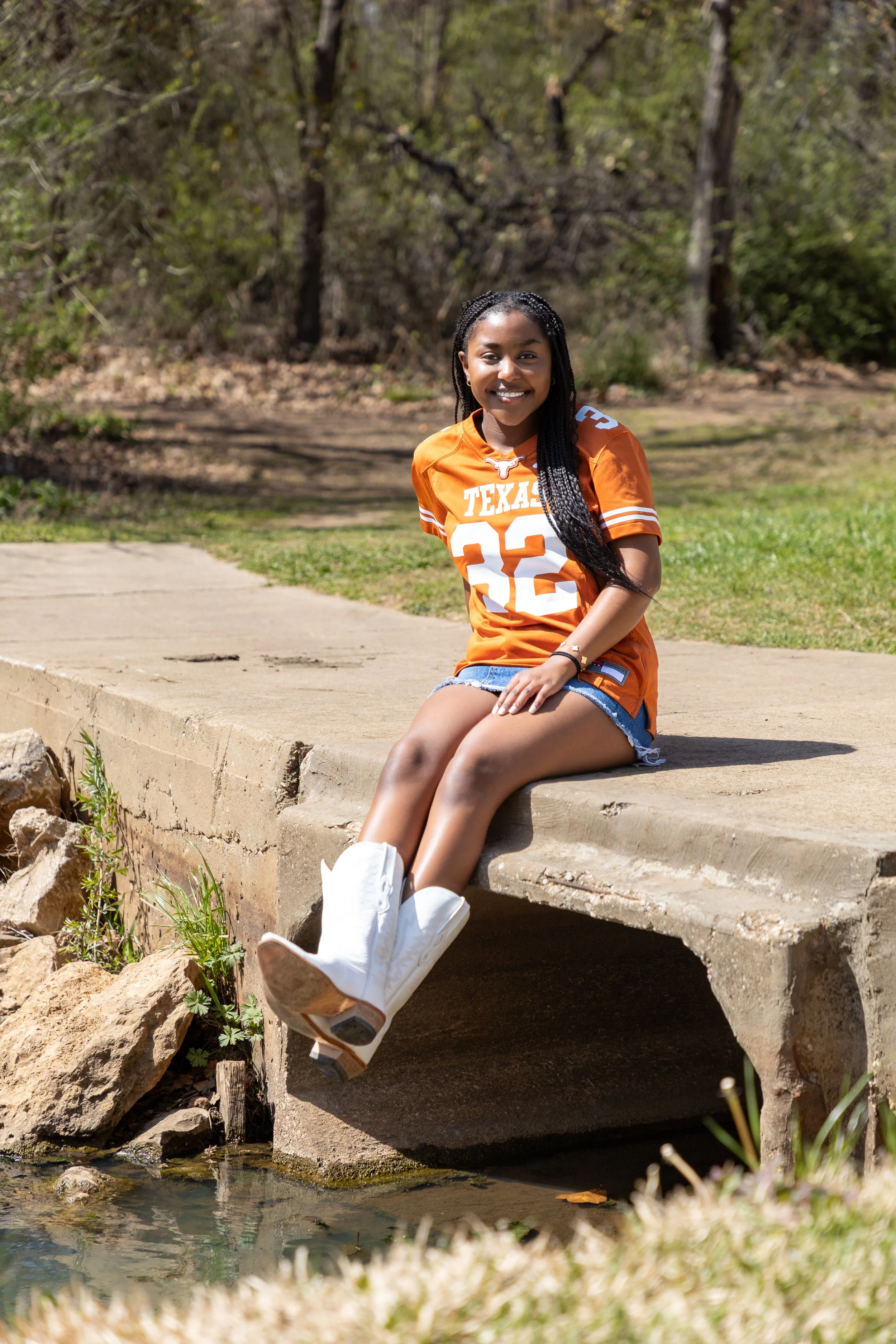 A young woman in a Texas Longhorns football jersey and denim shorts sitting on a concrete ledge by a small creek, smiling at the camera outdoors.