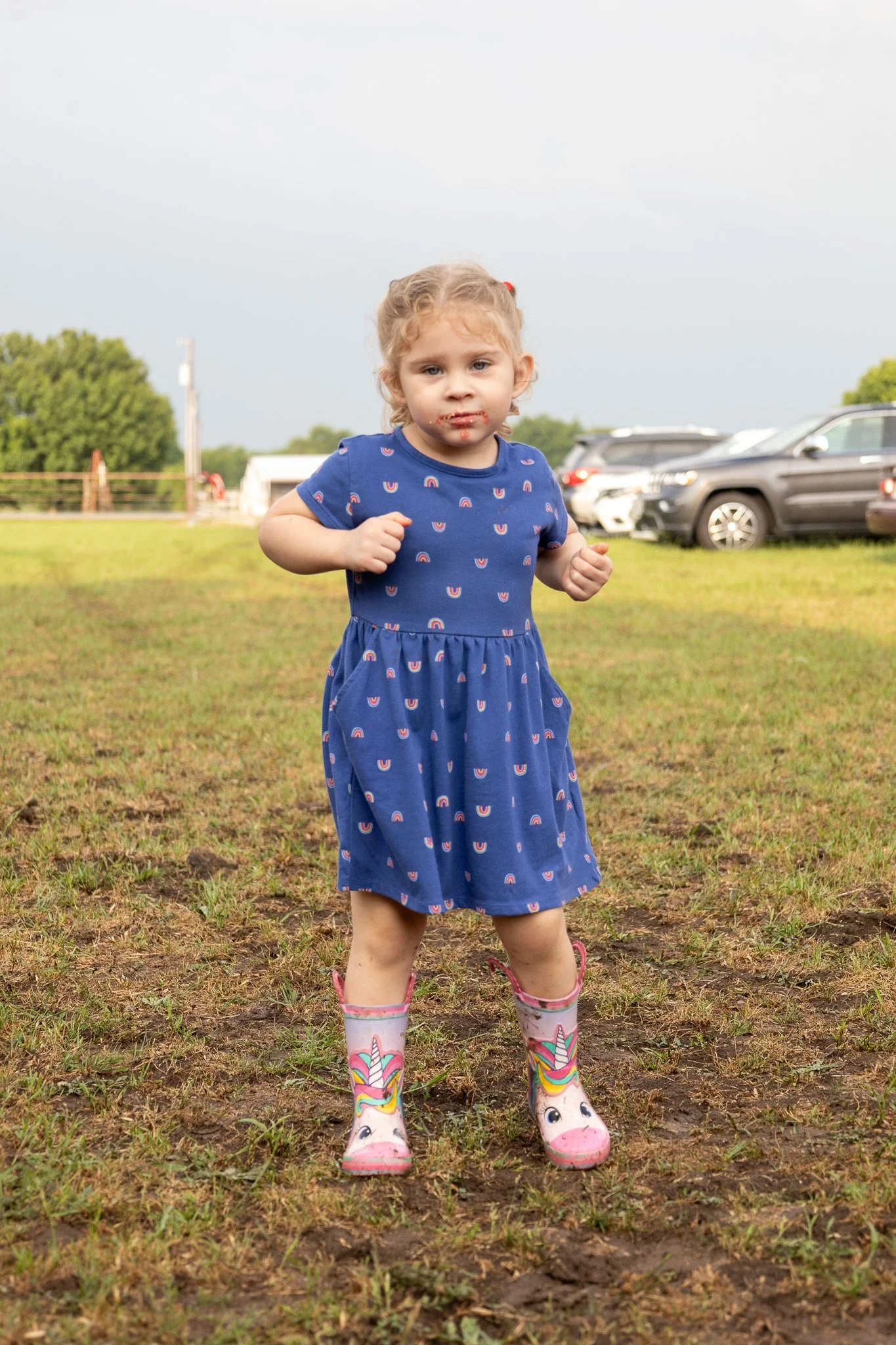 A young girl in a blue dress with a rainbow pattern, wearing unicorn rain boots, running outdoors on a grassy field with dirt patches, parked cars, and trees in the background under a cloudy sky.