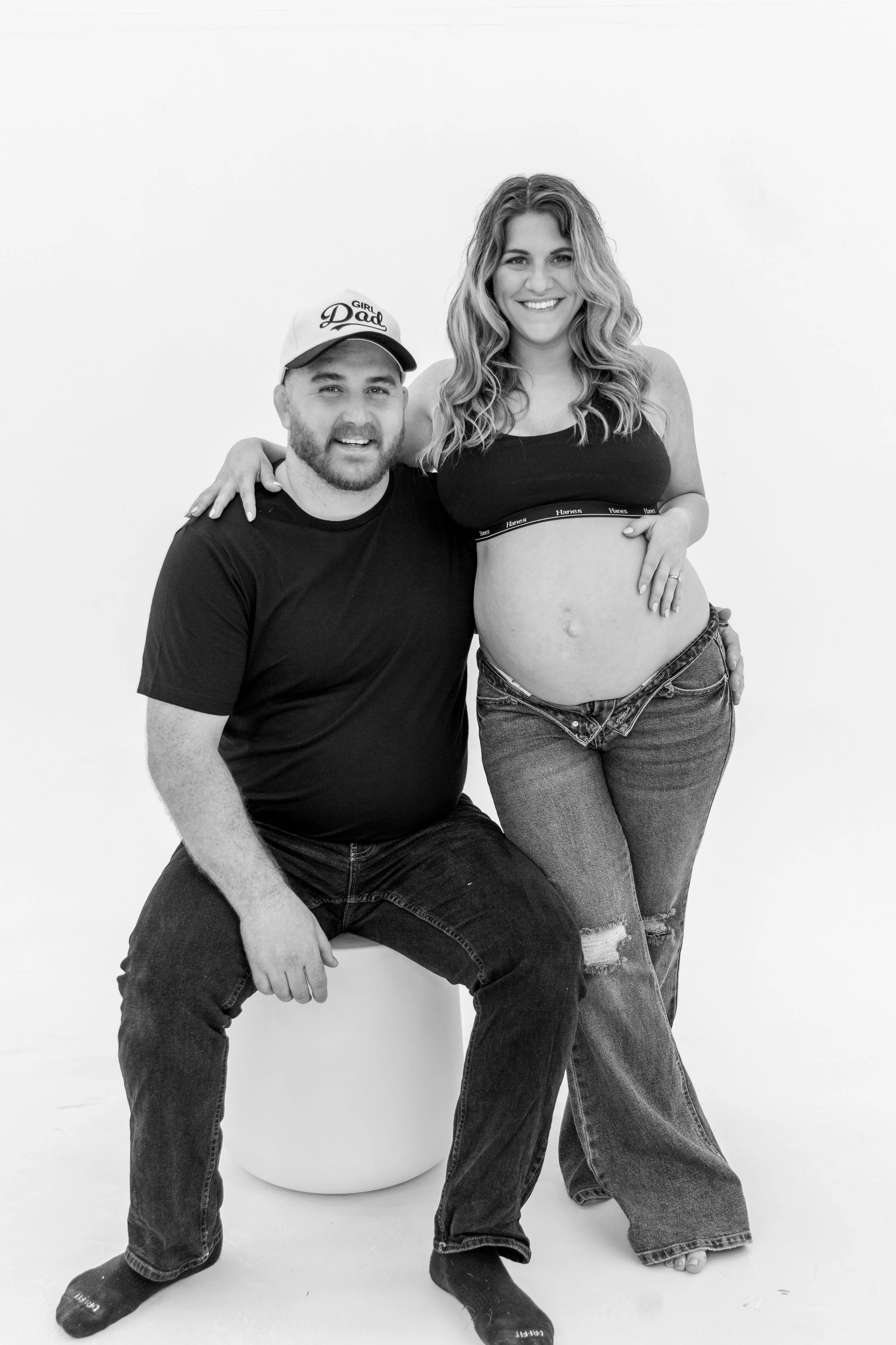 A man and a woman posing together in a studio with a plain white background. The man is wearing a cap that says 'Girl Dad' and a black t-shirt. The woman is pregnant, wearing a sports bra and jeans, and has her arm around the man's shoulder. Both are