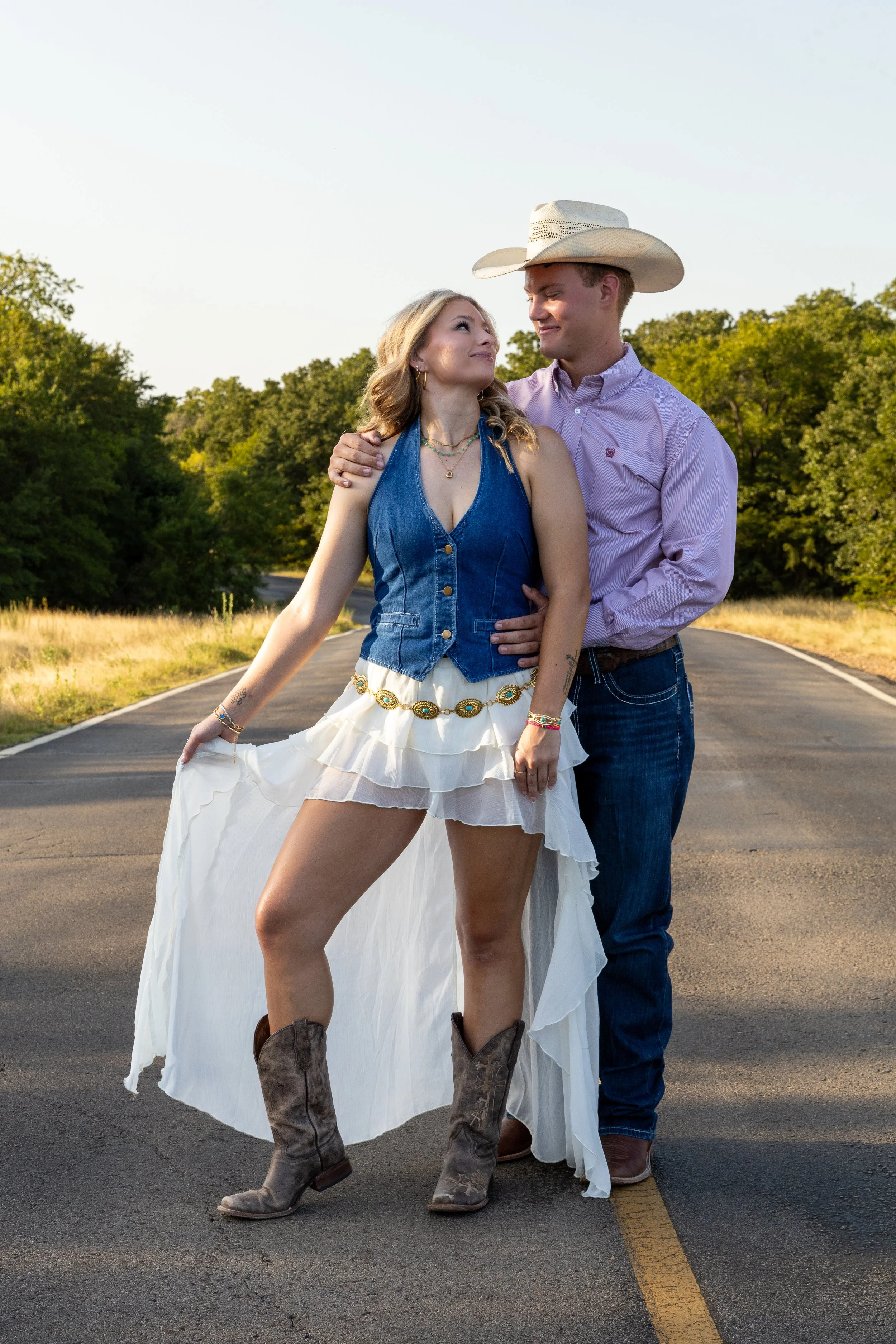 A young woman in a white dress, denim vest, and cowboy boots posing with a young man in a button-up shirt, jeans, and a cowboy hat on an empty rural road with green trees in the background.