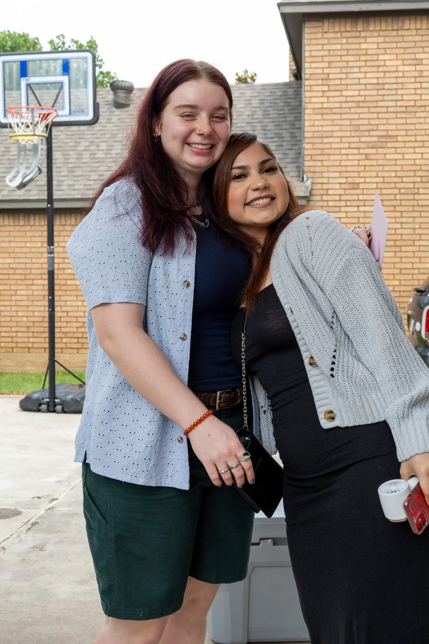Two young women smiling and hugging outdoors in a residential driveway, with a basketball hoop and brick house in the background.