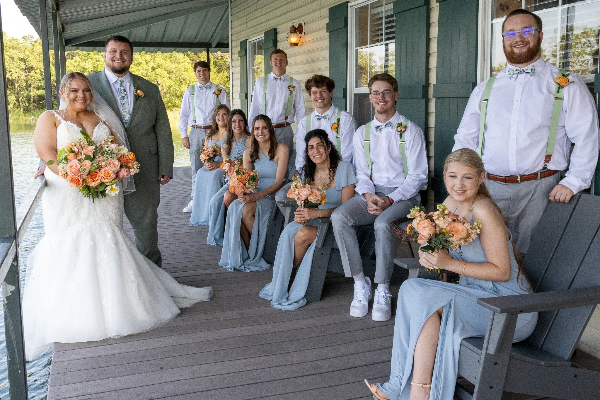 Group of bridesmaids and groomsmen sitting and standing on a porch near a lake, with a bride and groom in the front left, all dressed in wedding attire and holding bouquets of flowers.