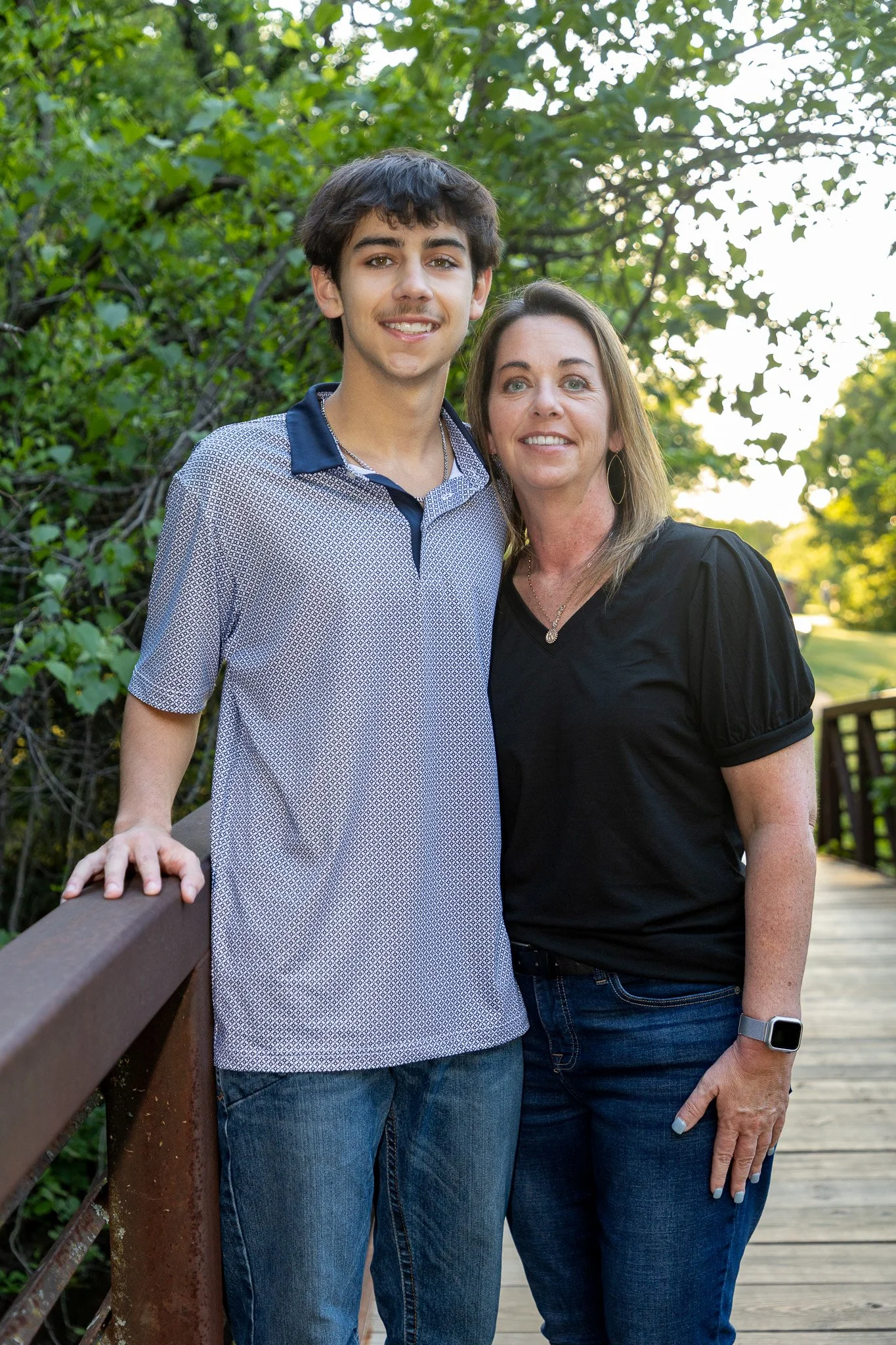 A teenage boy and a woman standing on a wooden bridge outdoors, smiling, surrounded by green trees in the evening sunlight.