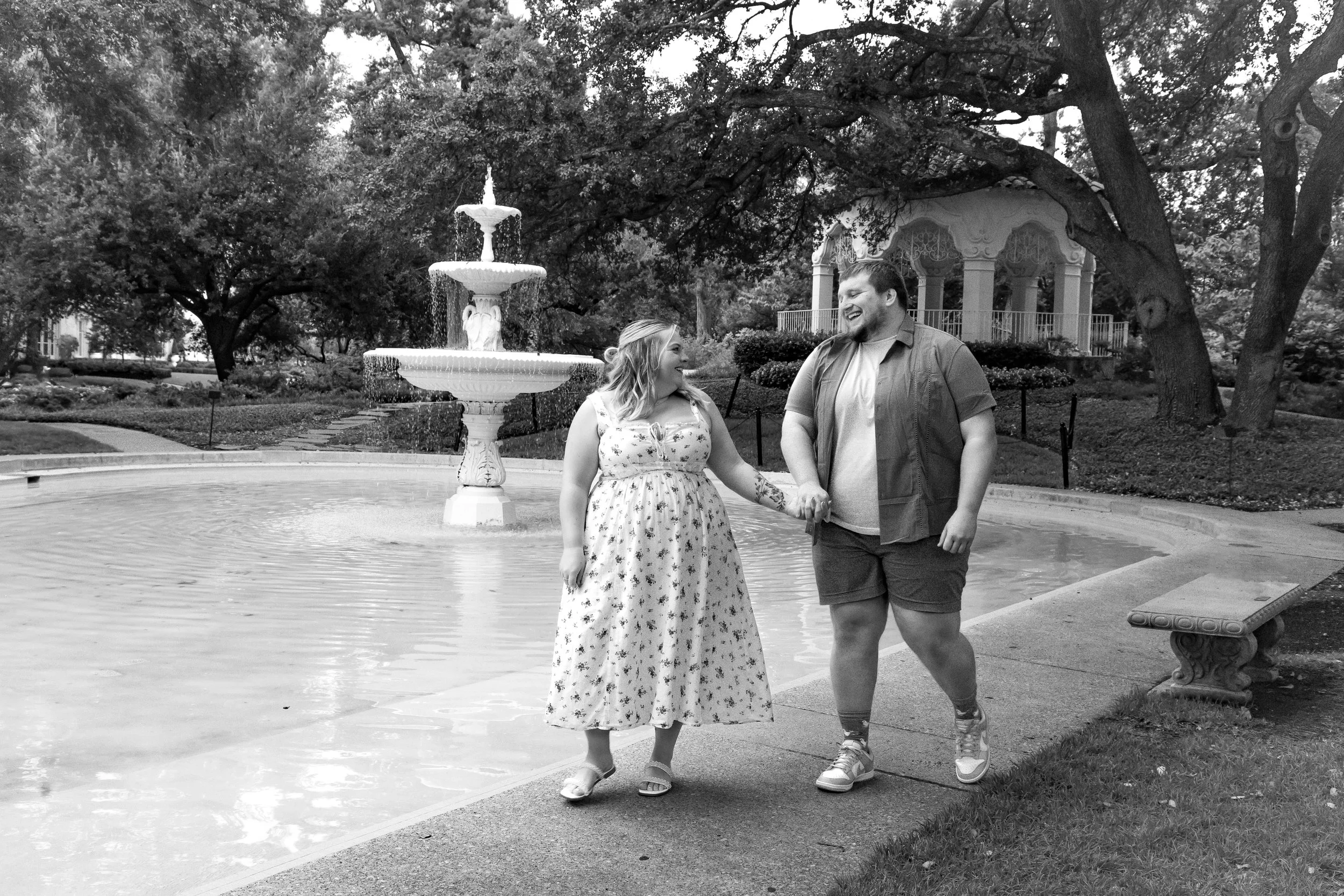 A smiling couple walks hand-in-hand near a fountain in a park, surrounded by trees and a gazebo in the background.