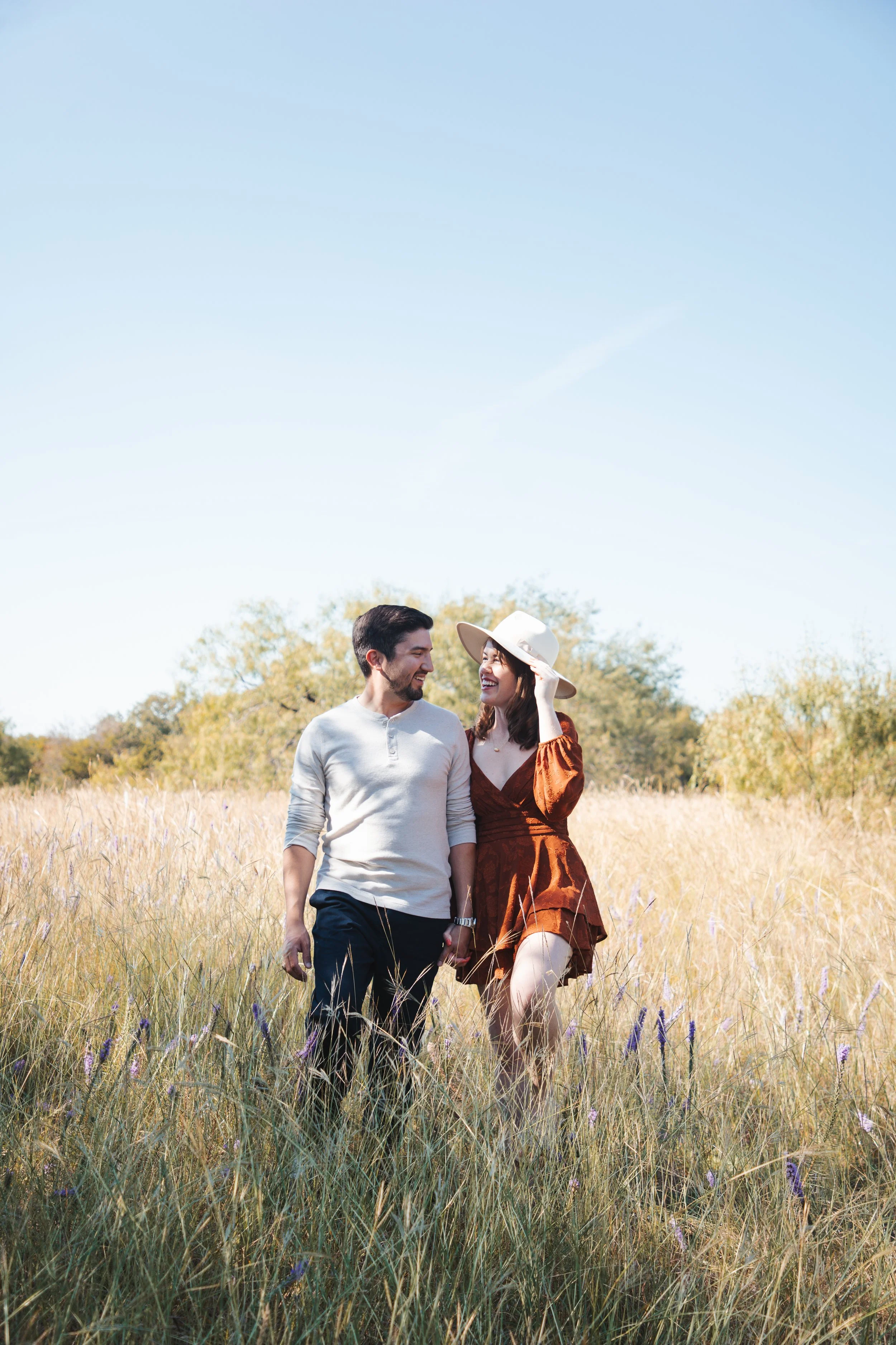 A man and woman walking together through a field of tall grass, smiling at each other, with the woman wearing a wide-brimmed hat and a rust-colored dress, and the man in a light gray shirt and dark pants.