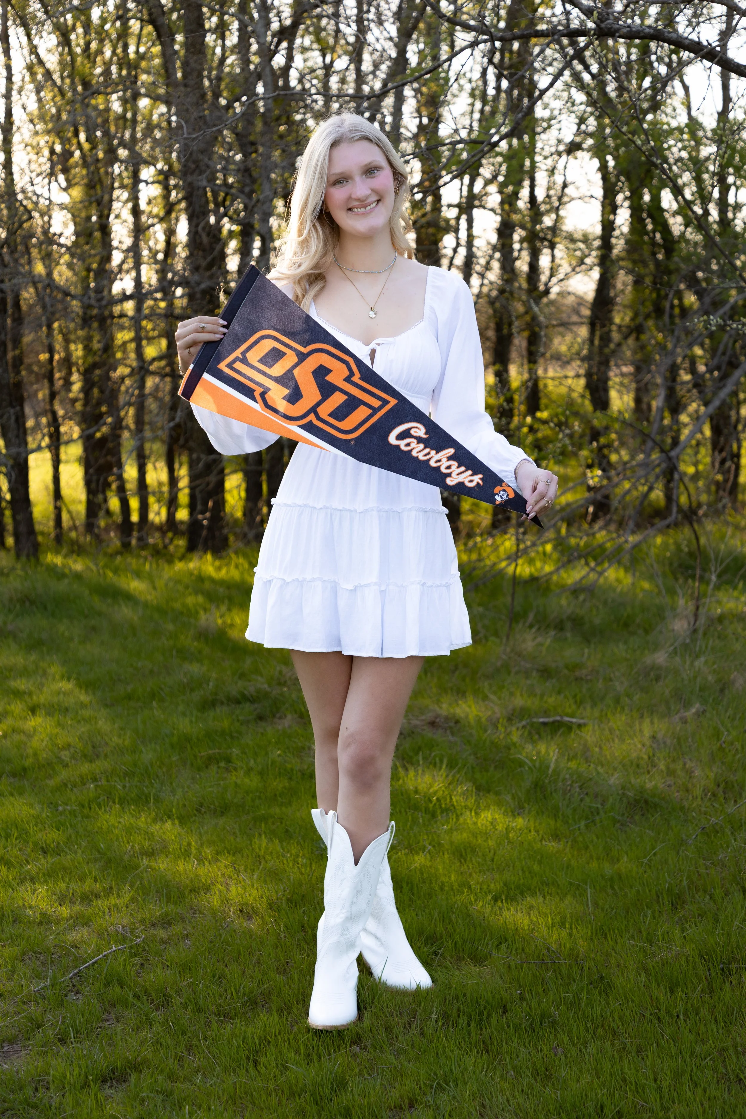 Young woman in a white dress and cowboy boots holding an Oklahoma State University Cowboys pennant outdoors in a grassy area with trees in the background.