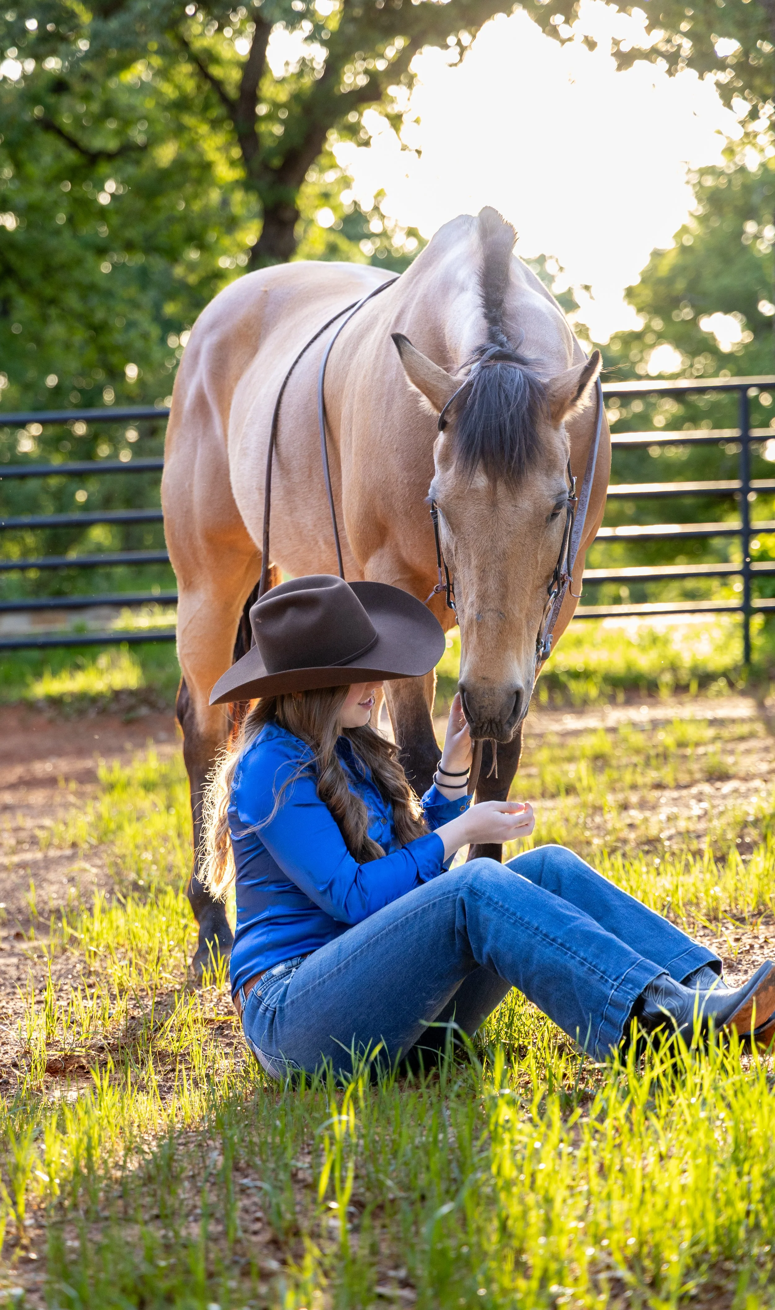 A young woman sitting on the grass in a blue shirt and jeans, wearing a large cowboy hat, gently petting a light-colored horse with a dark mane in a fenced outdoor area during sunlight.