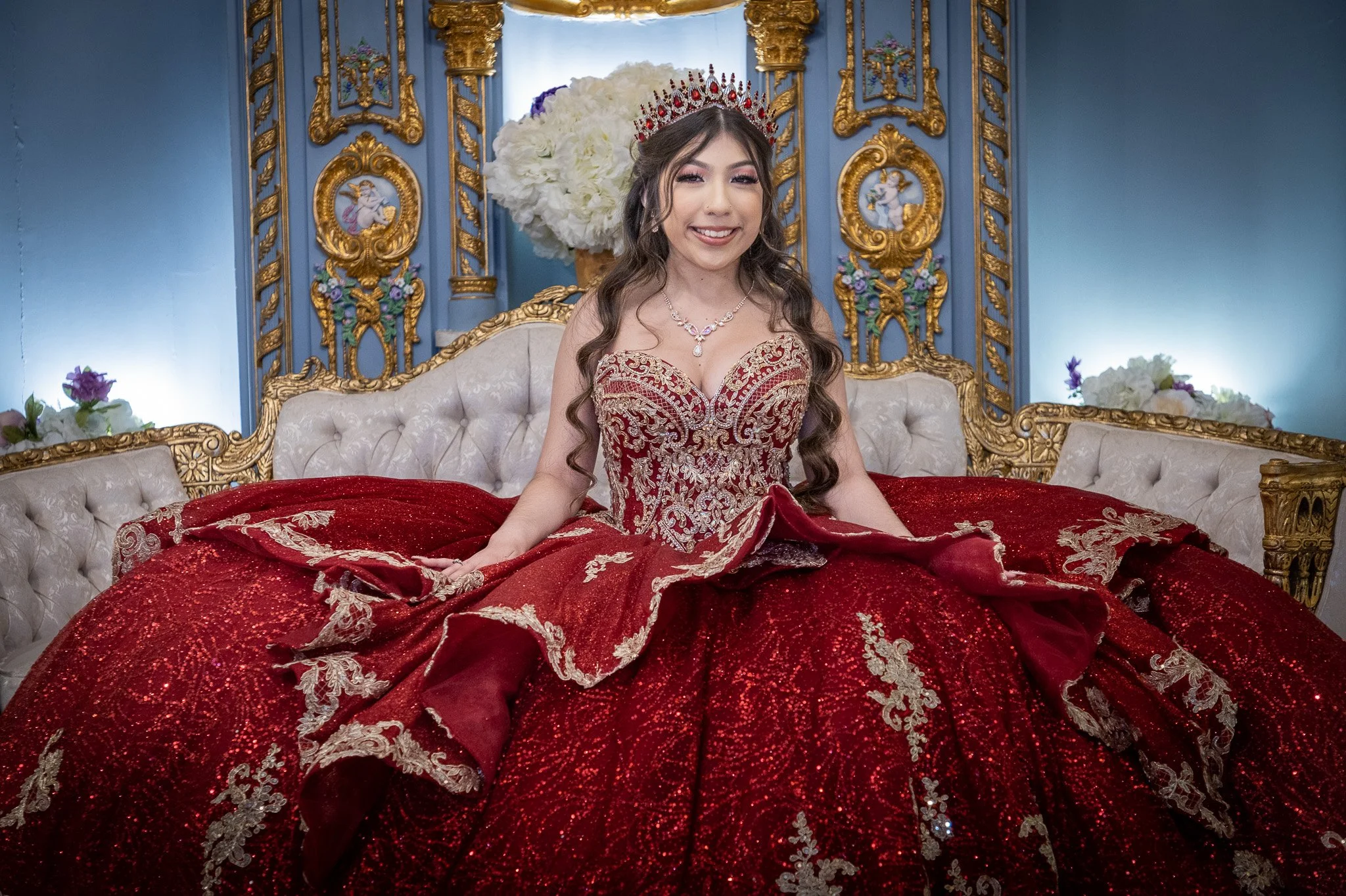 A woman wearing a red and gold ornate gown, sitting on an elegant beige and gold couch in a decorated room with blue walls, gold accents, and floral arrangements behind her.
