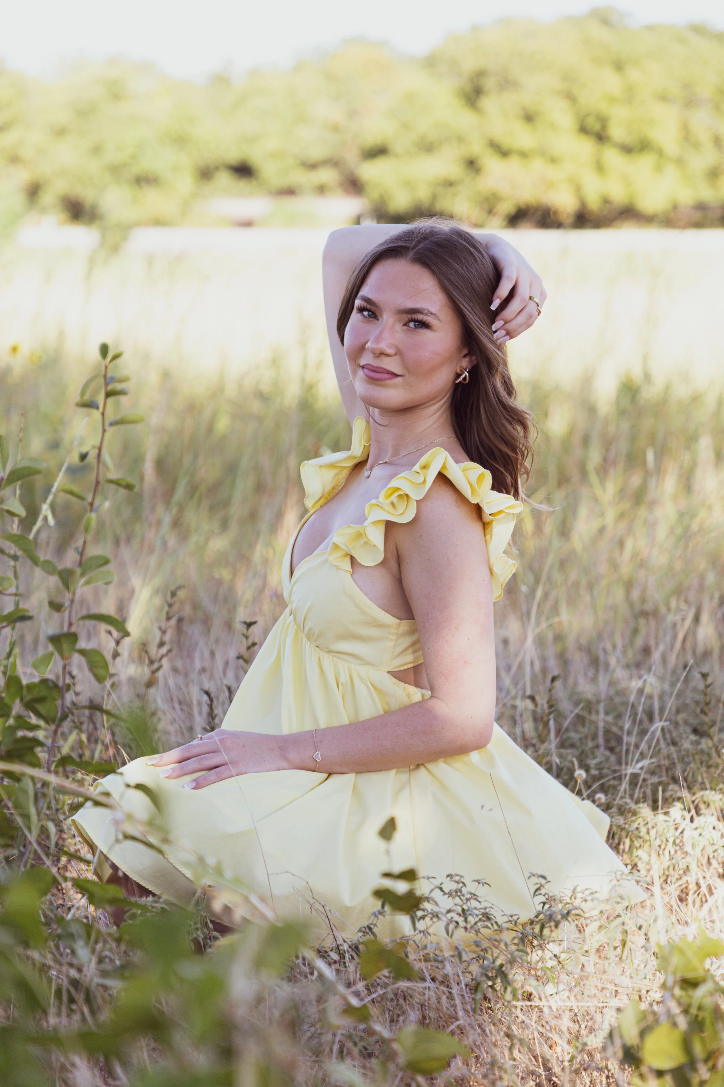 A young woman in a yellow dress sitting on the ground in a grassy field with trees in the background.