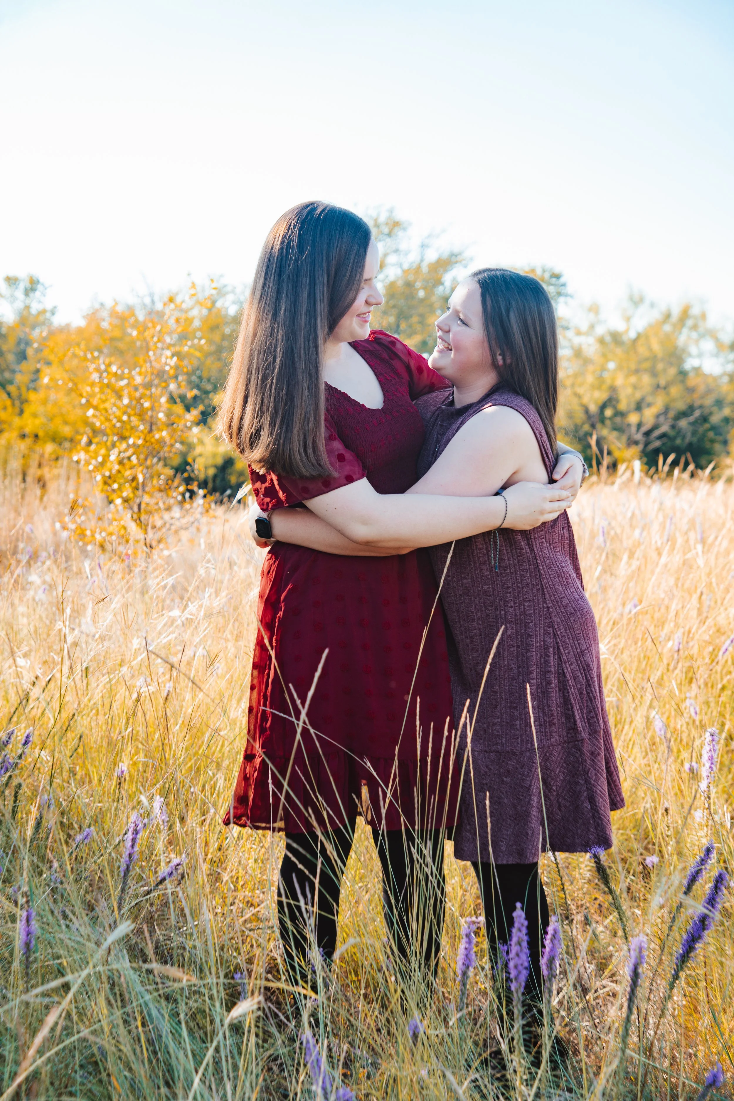 Two women hugging in a field with fall foliage, wearing burgundy dresses.