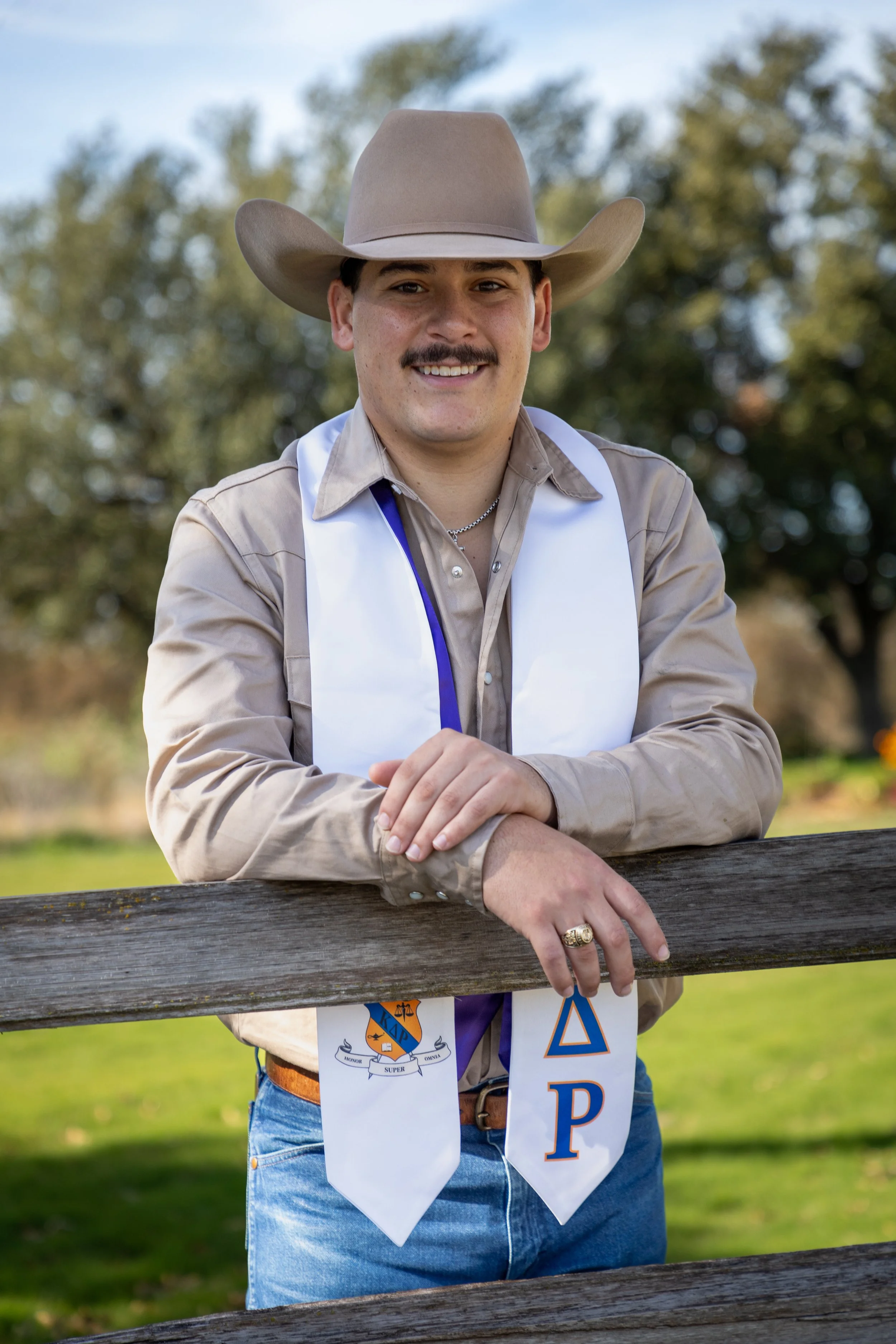 A young man wearing a cowboy hat and a white sash with Greek letters standing outdoors behind a wooden fence. He is wearing a beige shirt, blue jeans, and a gold ring.