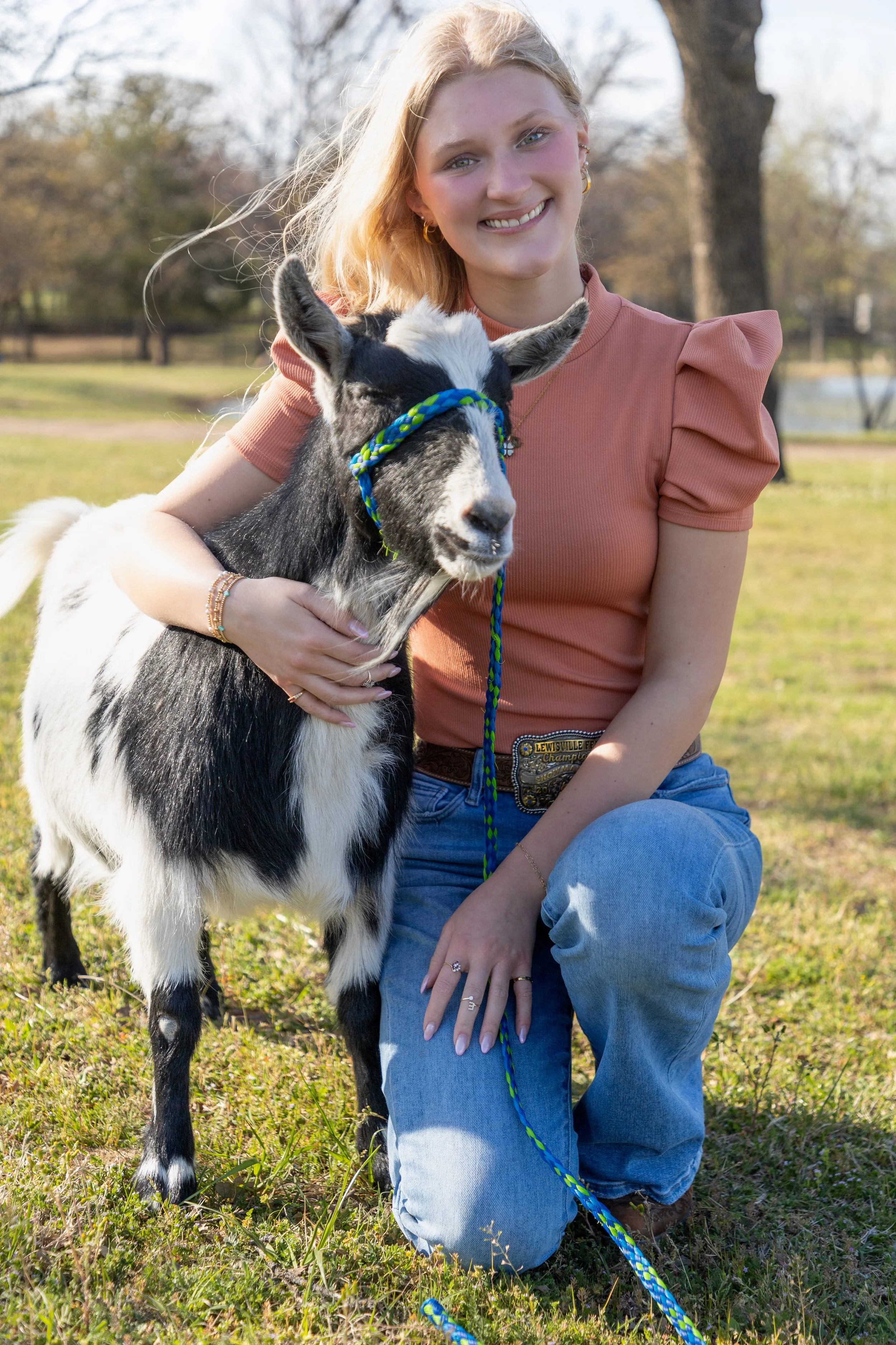A young woman with blonde hair, wearing a peach-colored top and blue jeans, kneels outdoors in a grassy area, smiling while holding a black and white baby goat with a colorful rope around its head.