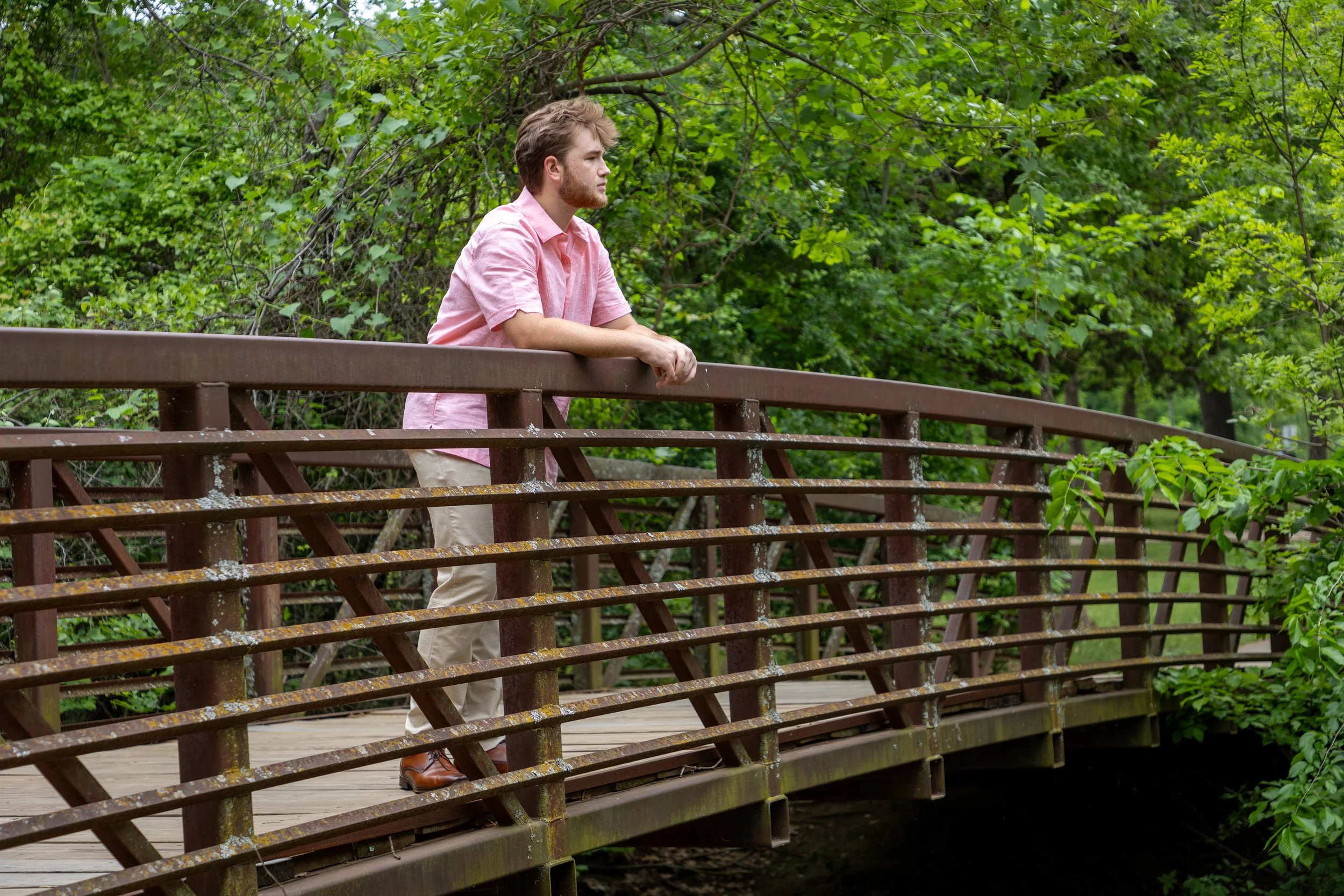 A young man with light brown hair and a beard, wearing a pink short-sleeved shirt and khaki pants, leaning on a wooden bridge railing and looking into the distance, surrounded by lush green trees.