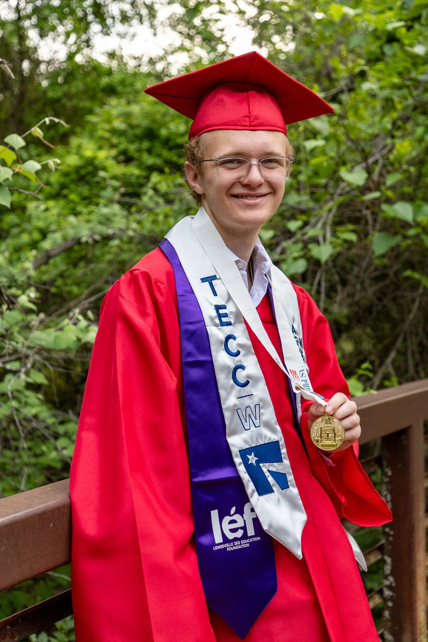 A young man in a red graduation gown and cap holding a gold medal, standing outdoors on a wooden bridge with green foliage in the background.