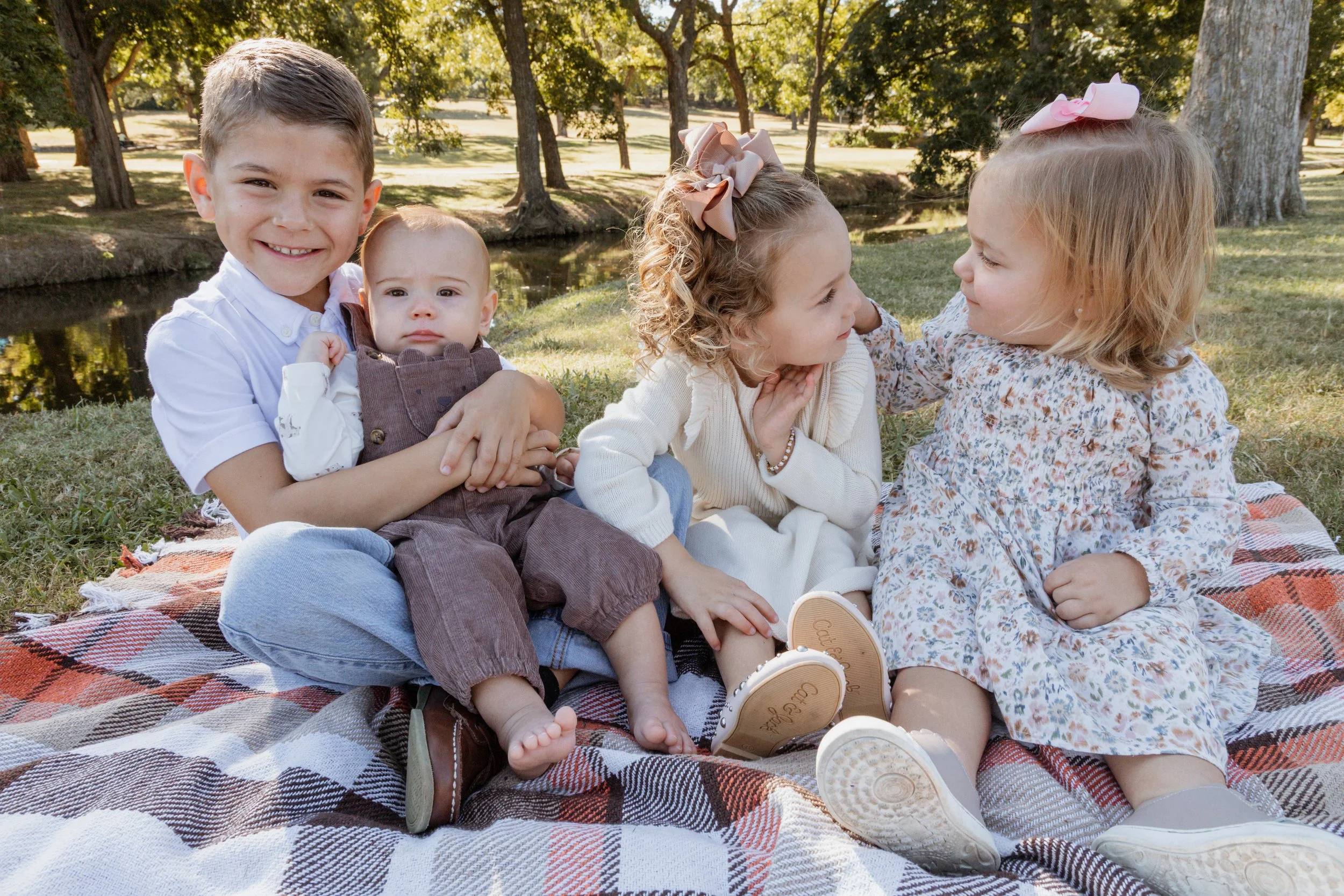 Four children sitting on a blanket by a pond in a park, engaging in conversation and smiling