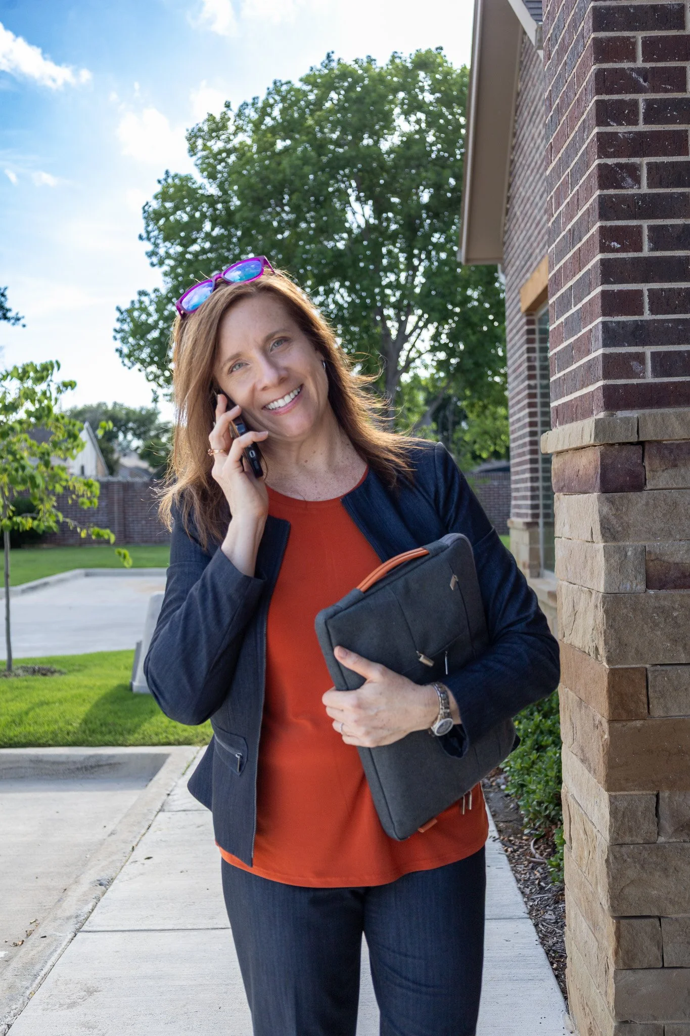 A woman with auburn hair, wearing sunglasses on her head, a dark blazer, and a burnt orange blouse, smiling while talking on her cellphone outside near a brick building, holding a black laptop case.