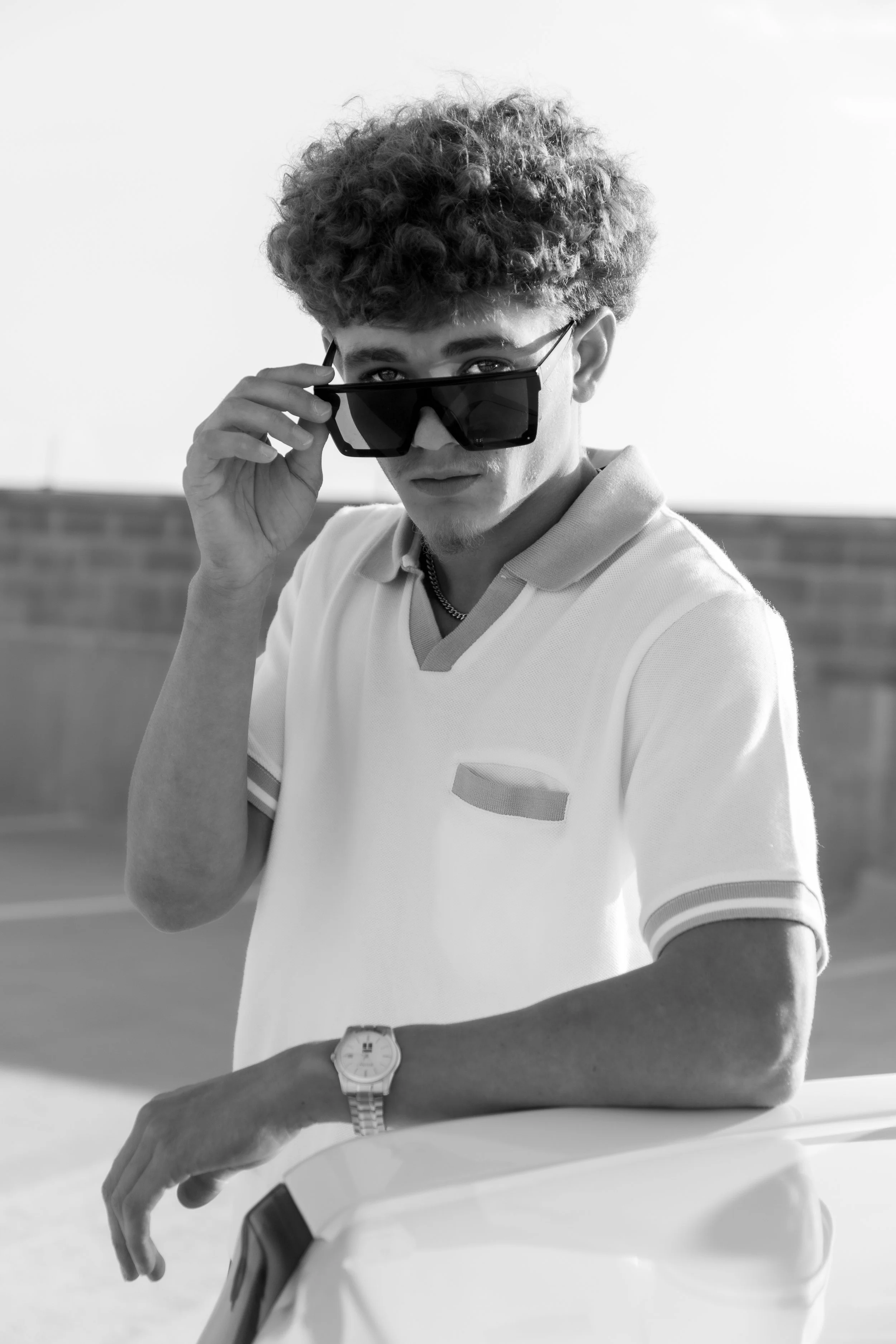 A young man with curly hair wearing sunglasses, a polo shirt, a watch, and a chain necklace, leaning on the hood of a car in an outdoor setting.