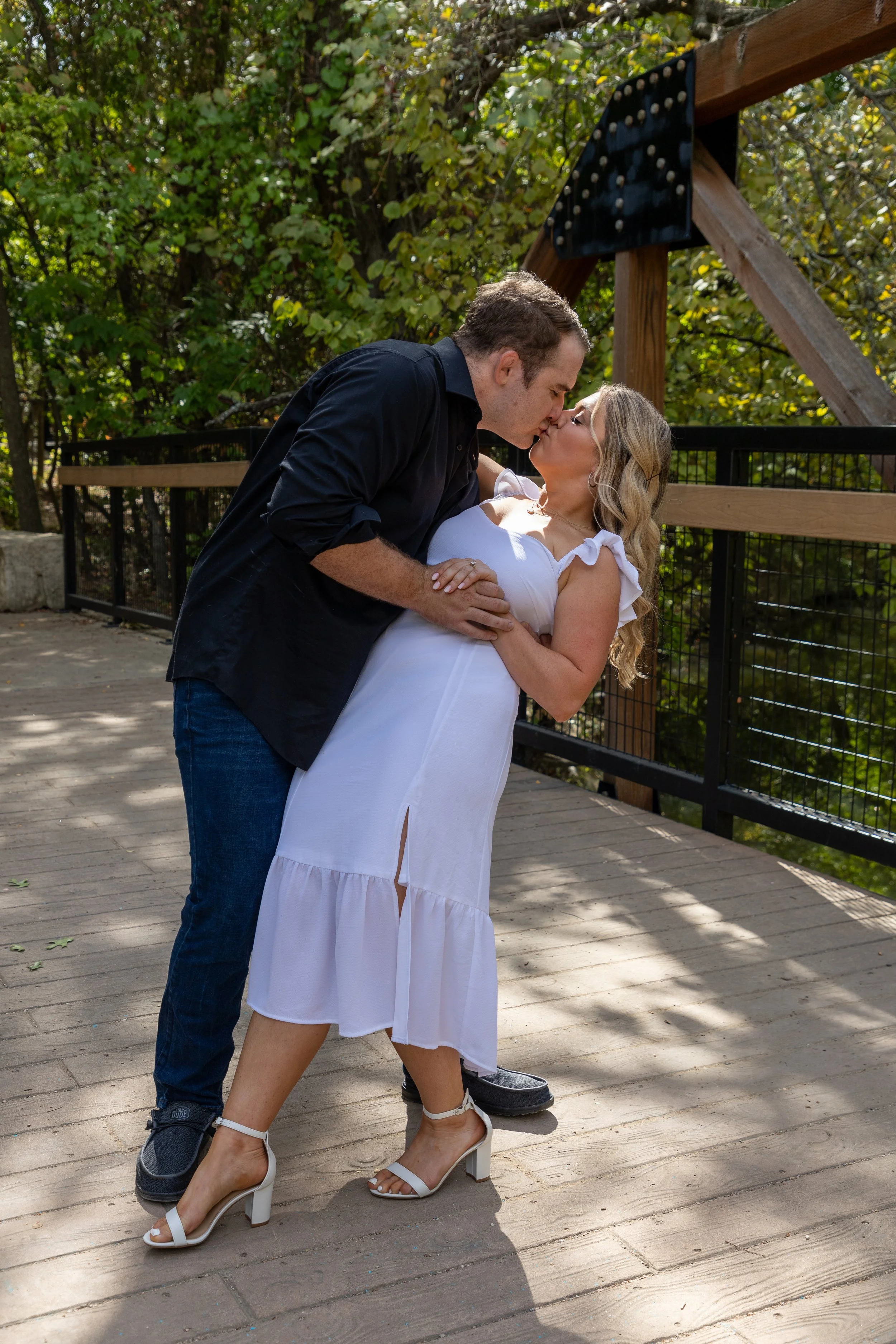 A couple sharing a kiss on a wooden deck surrounded by autumn trees. The woman in a white dress and heels stands while the man in a black shirt and jeans leans in for a kiss.