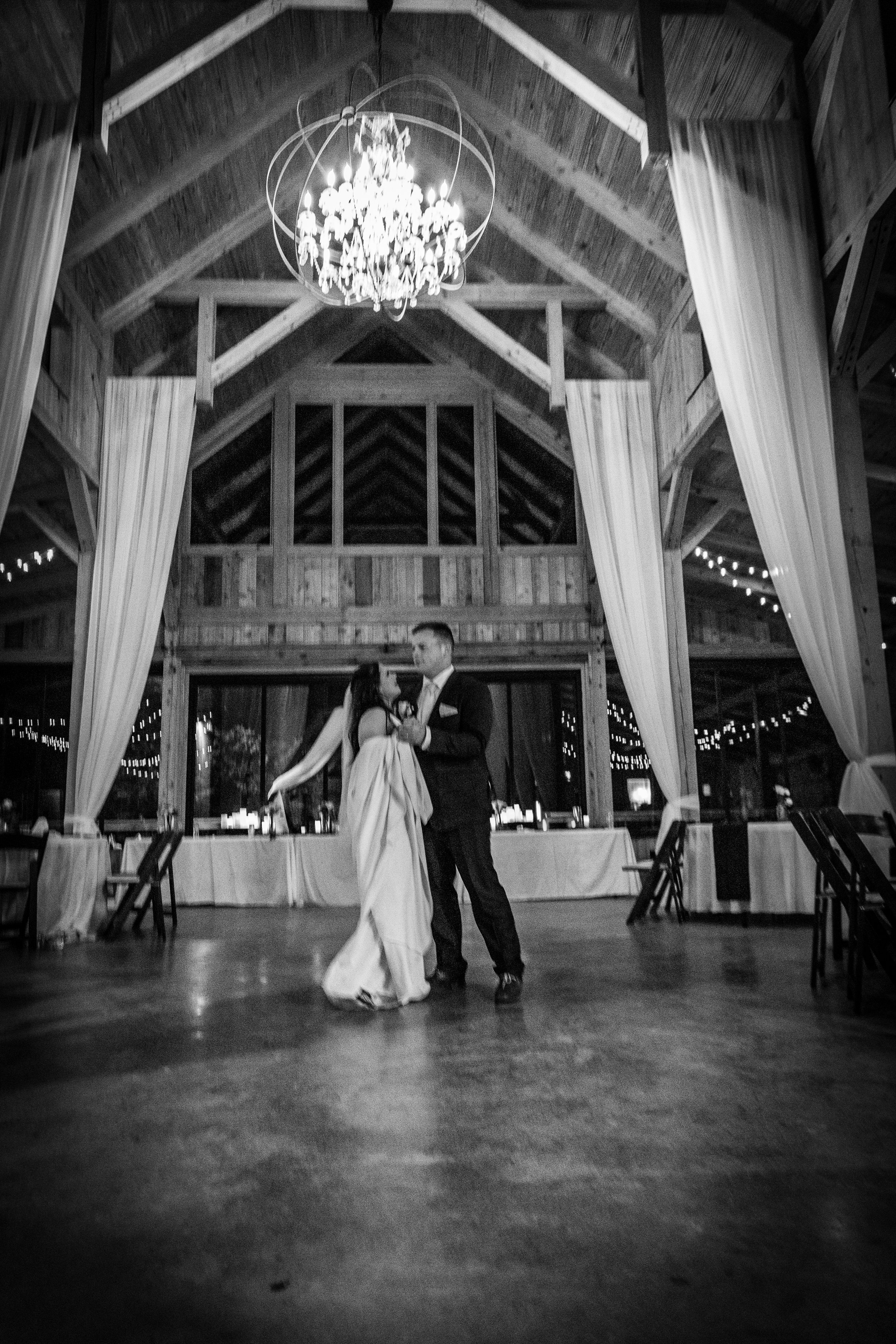 A bride and groom dancing in a rustic wood wedding venue decorated with white curtains and string lights, with a chandelier hanging from the ceiling.
