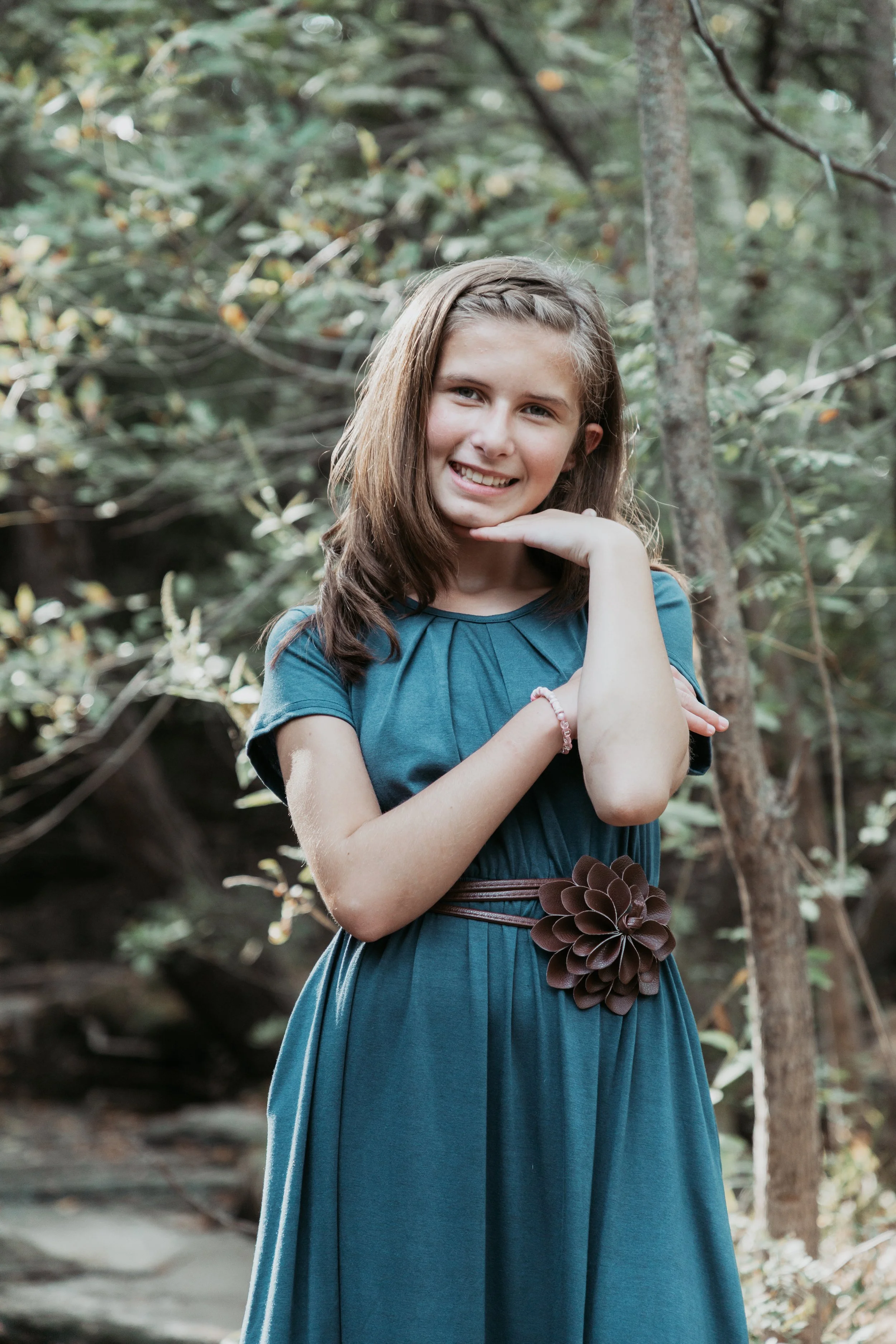 A young girl with shoulder-length brown hair, wearing a teal dress with a large flower belt, standing outdoors in a wooded area, smiling and posing with her hand under her chin.
