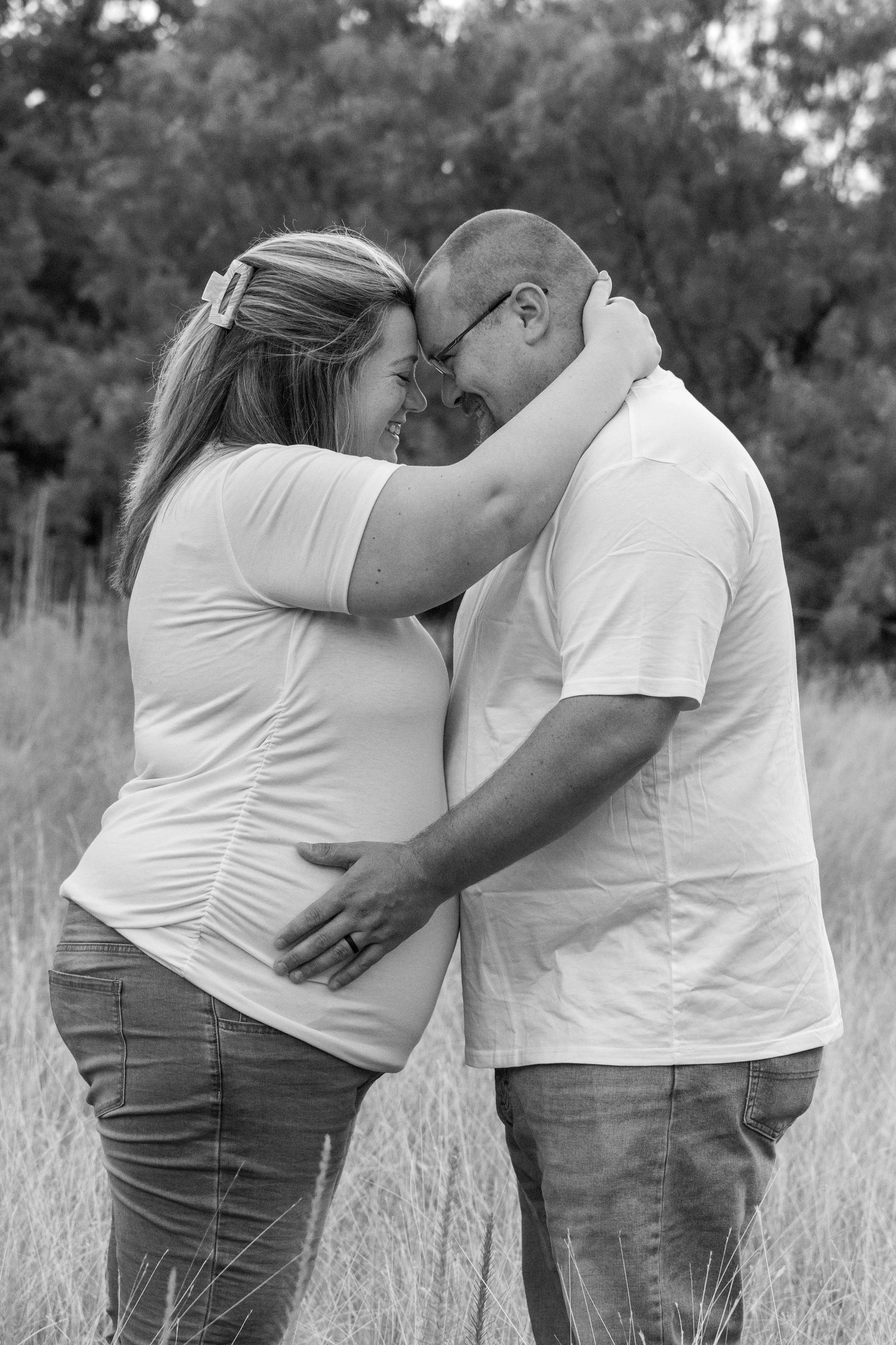 A pregnant woman and a man face each other in a field, touching foreheads and smiling, with their hands on the woman's pregnant belly.