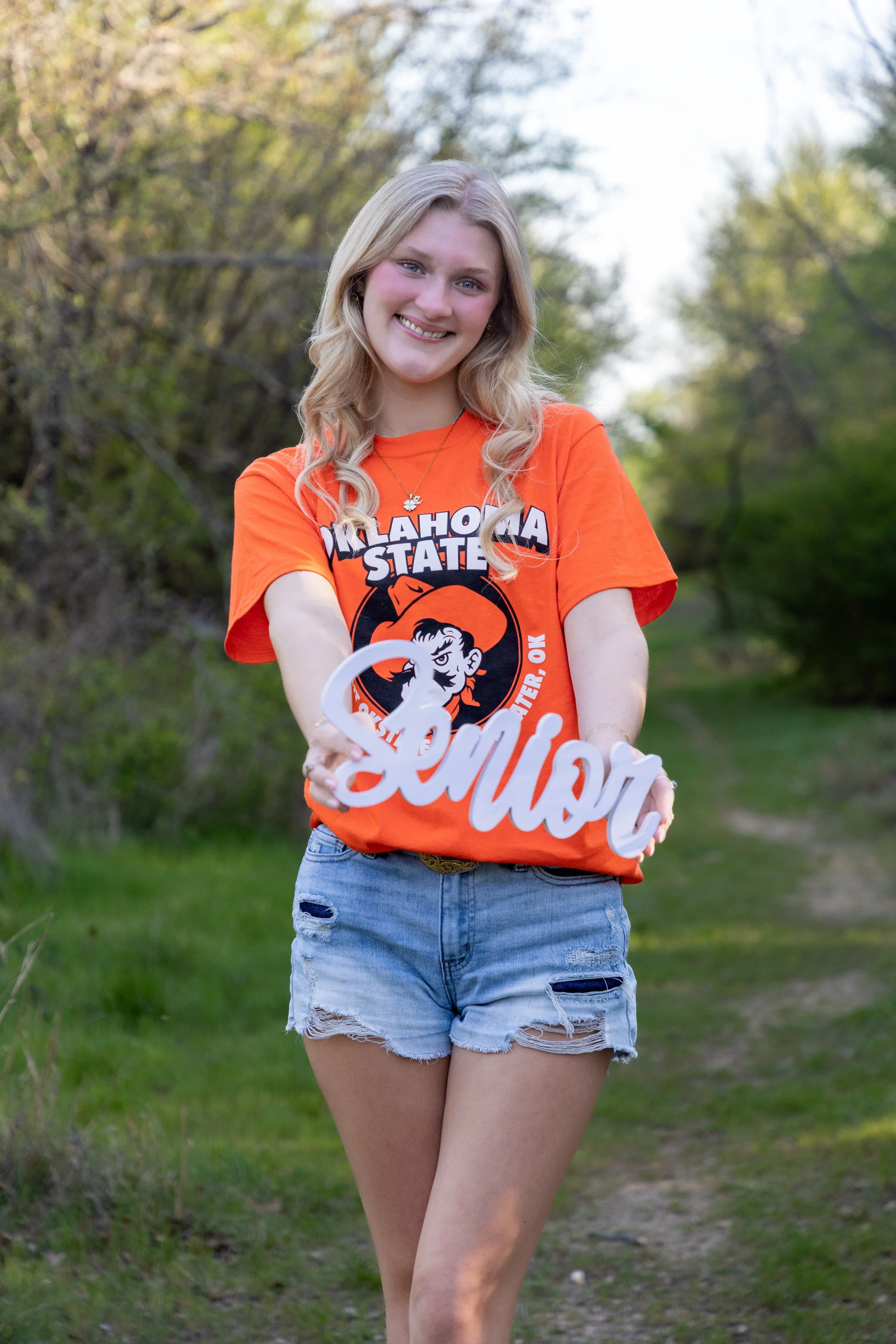 A young woman with blonde hair wearing an orange Oklahoma State shirt and ripped denim shorts, standing on a grassy trail and holding a white decorative sign that says 'Senior'.