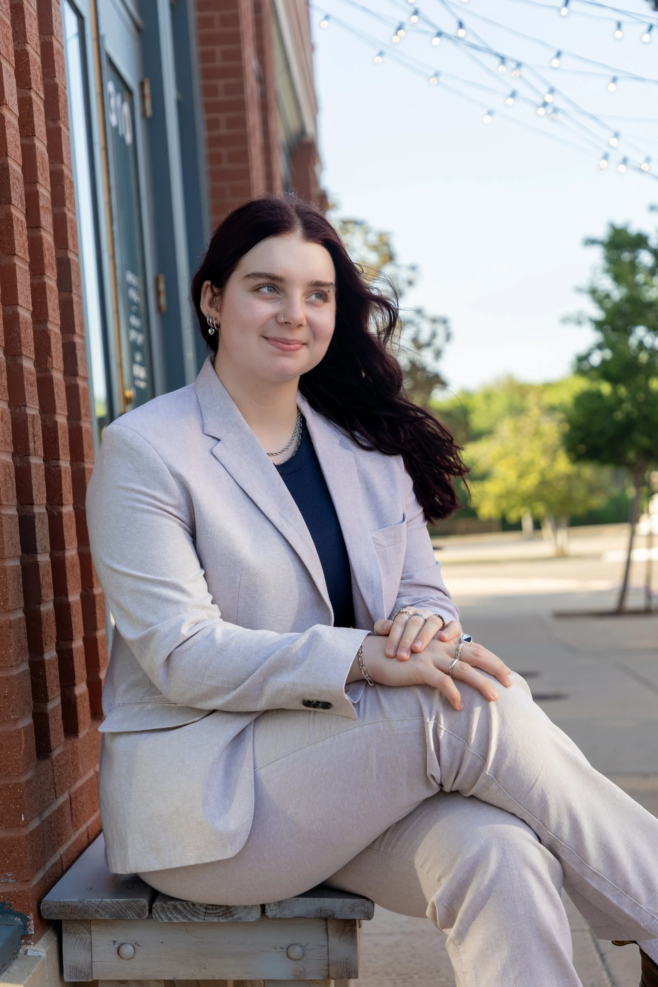 A young woman with dark hair and fair skin, wearing a light-colored suit and sitting on a bench outside on a sunny day, with string lights above and a brick building in the background.