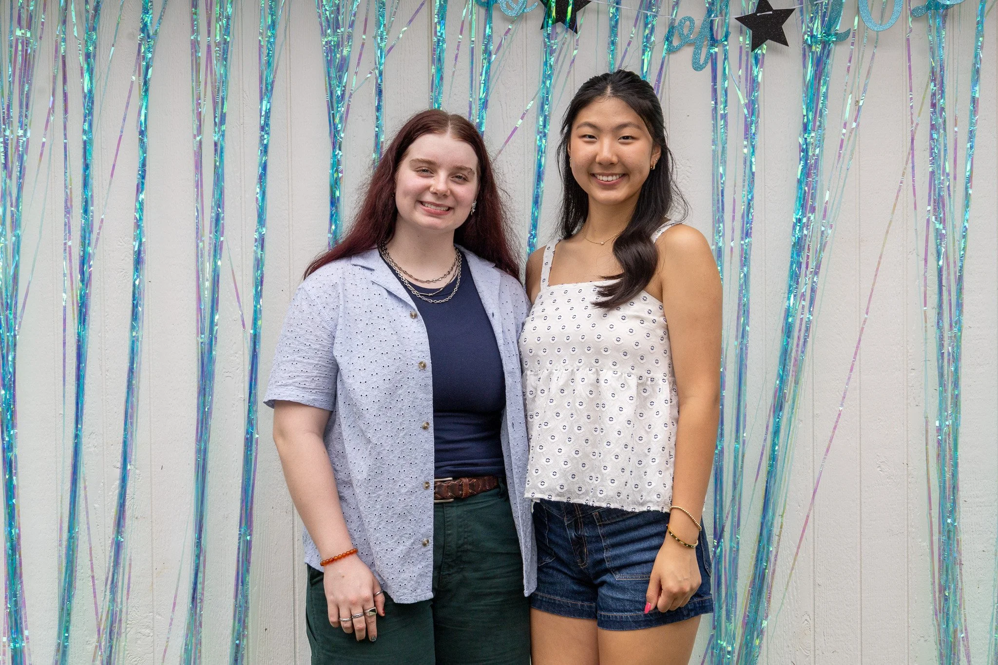 Two young women standing close together, smiling, in front of a light-colored wall with blue and purple streamers and a black star decoration, at a celebration or party.