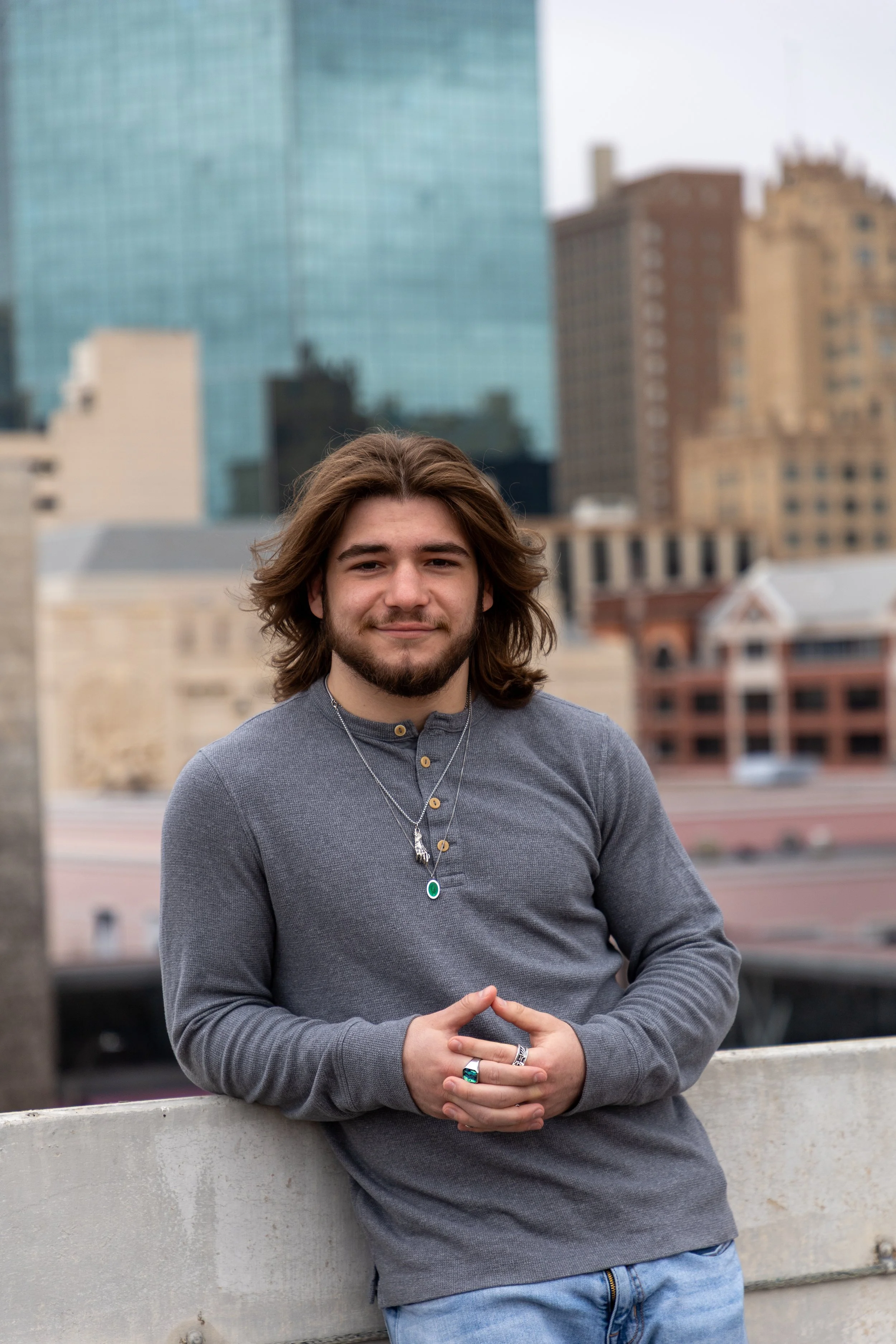 A young man with long hair and a beard standing outdoors on a rooftop with city buildings in the background.