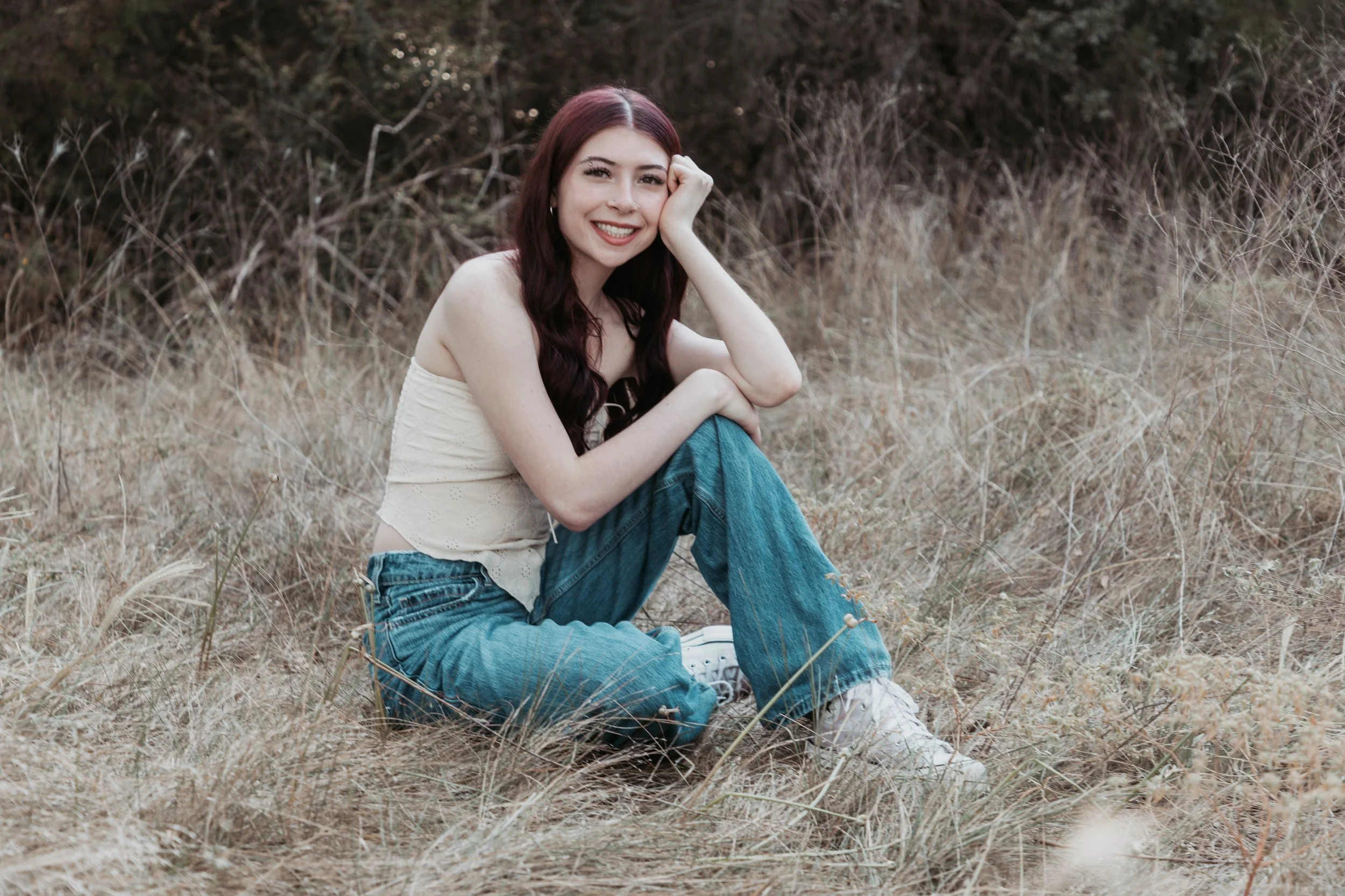 A young woman with long, wavy dark red hair sitting in dry grass outdoors, smiling at the camera, wearing a cream-colored sleeveless top, blue jeans, and white sneakers.