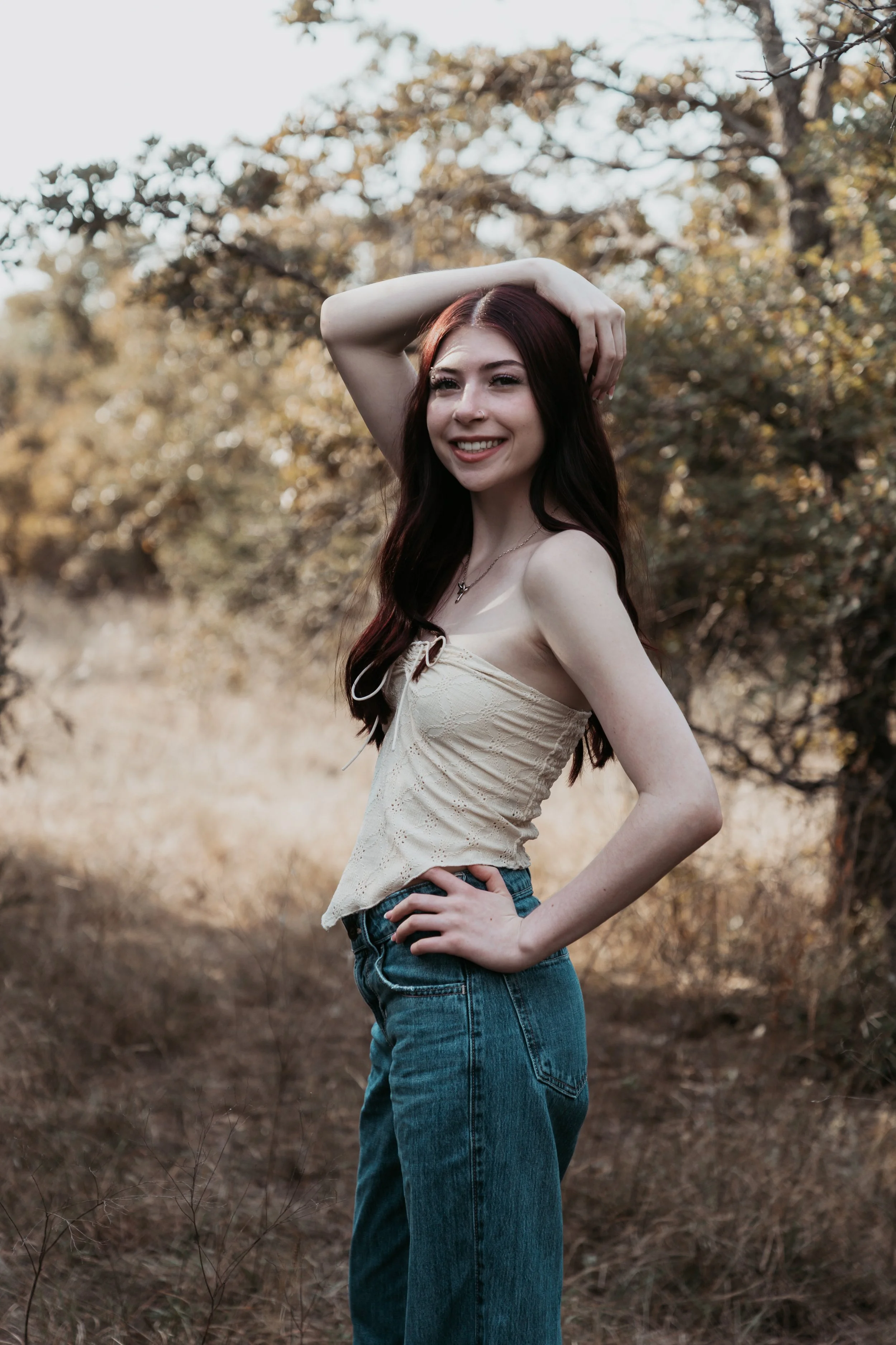 A young woman with long, dark red hair smiling, standing outdoors in a natural setting with trees and dry grass, wearing a light-colored sleeveless top and blue jeans.
