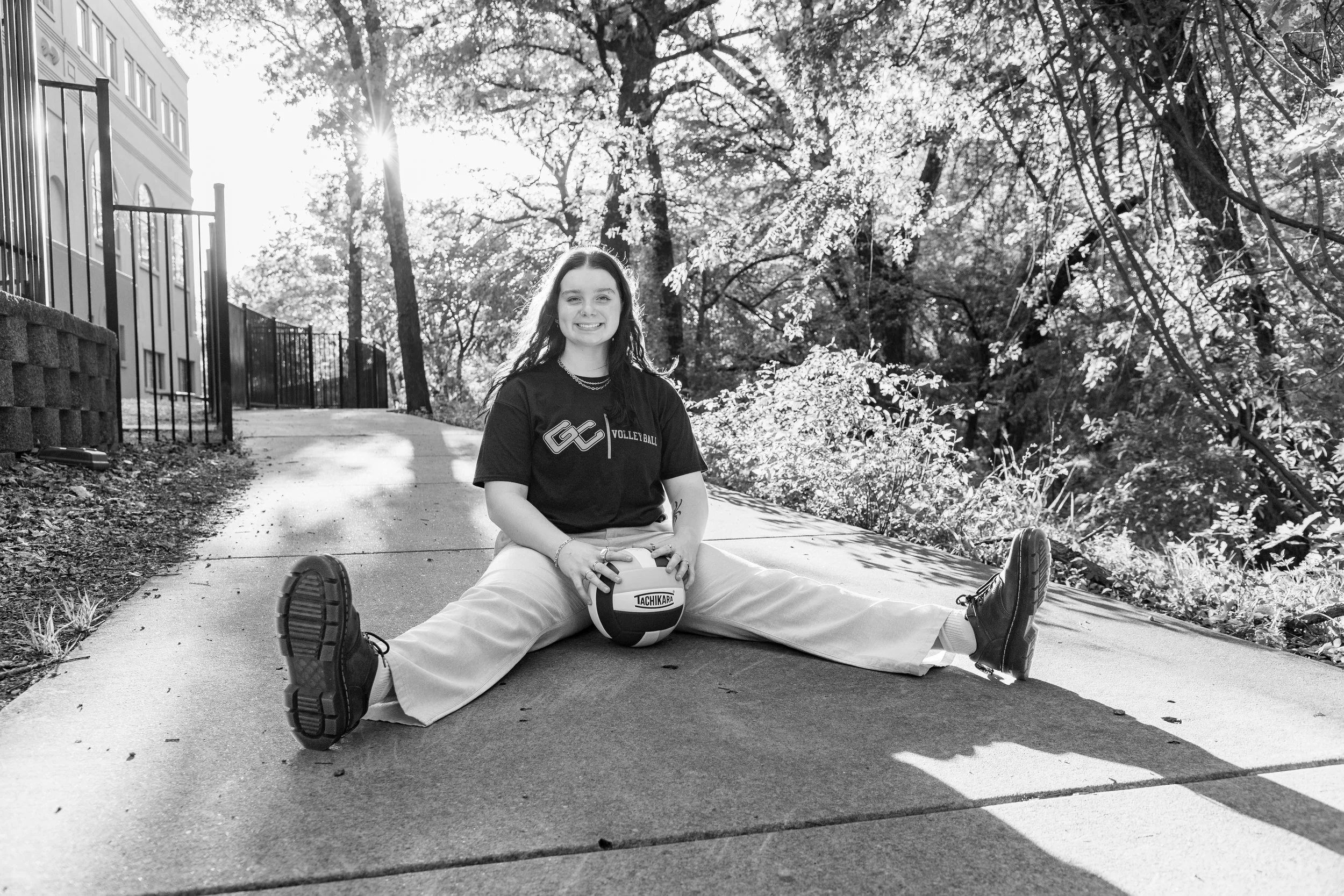 A young woman sitting on a sidewalk in a split with her legs extended and holding a volleyball, smiling at the camera, with sunlight filtering through trees in the background.