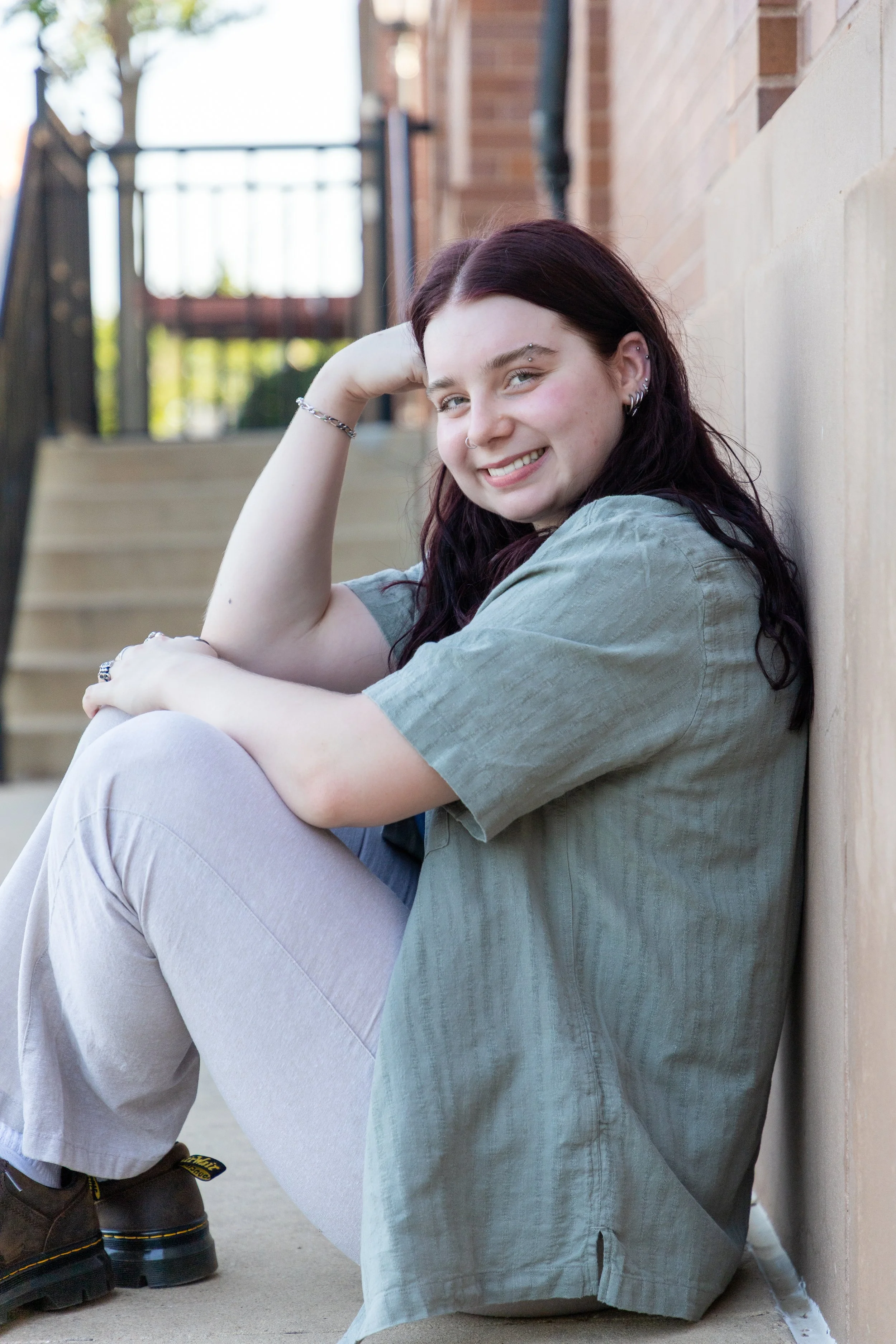 A young woman with dark hair and piercings, sitting on the ground next to a brick wall, smiling at the camera.