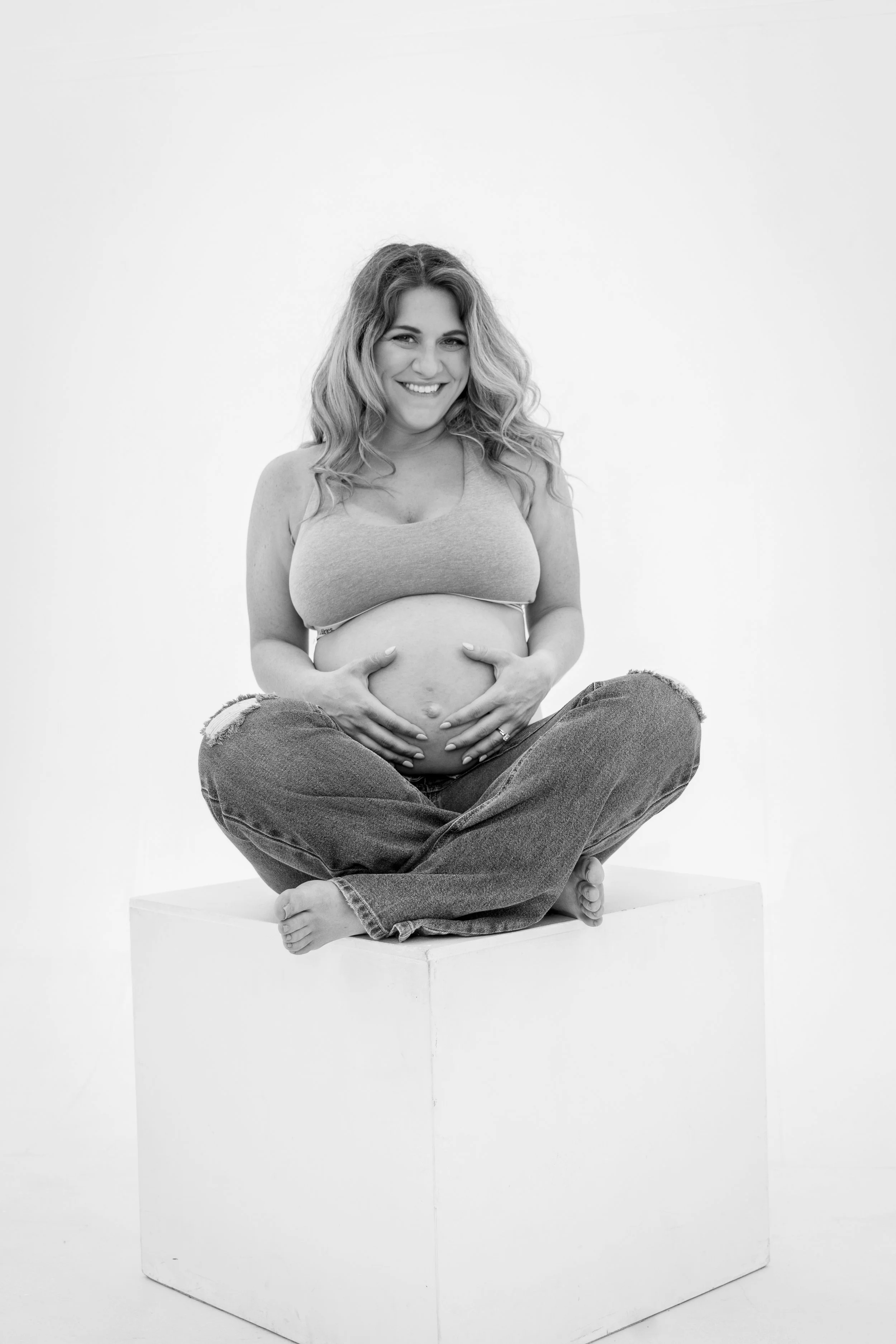 Black and white photo of a pregnant woman sitting cross-legged on a platform, smiling and holding her belly, with a plain white background.