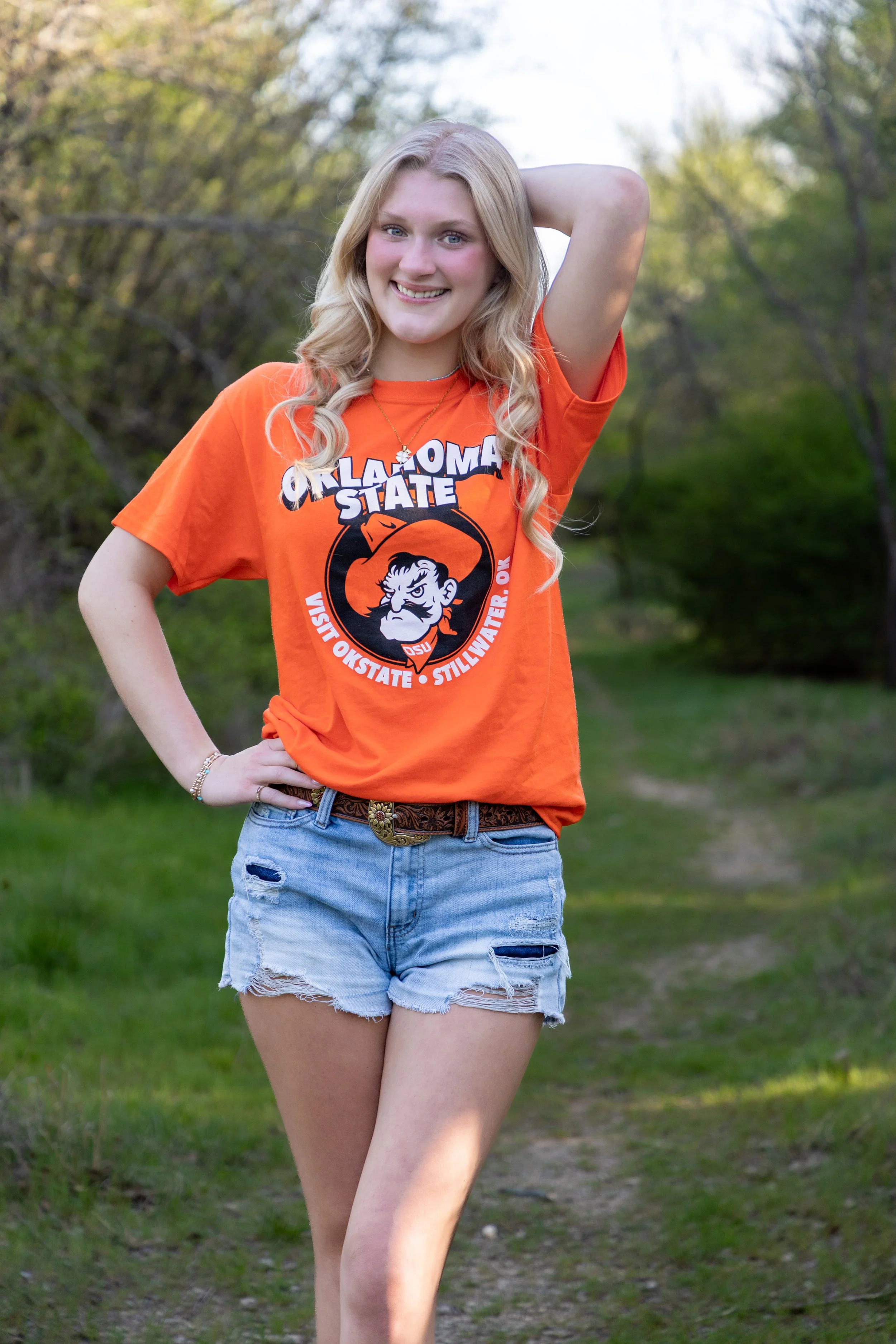 A young woman with blonde curly hair standing outdoors on a grassy trail, smiling with her hand on her hip, wearing an orange Oklahoma State University t-shirt and ripped denim shorts.